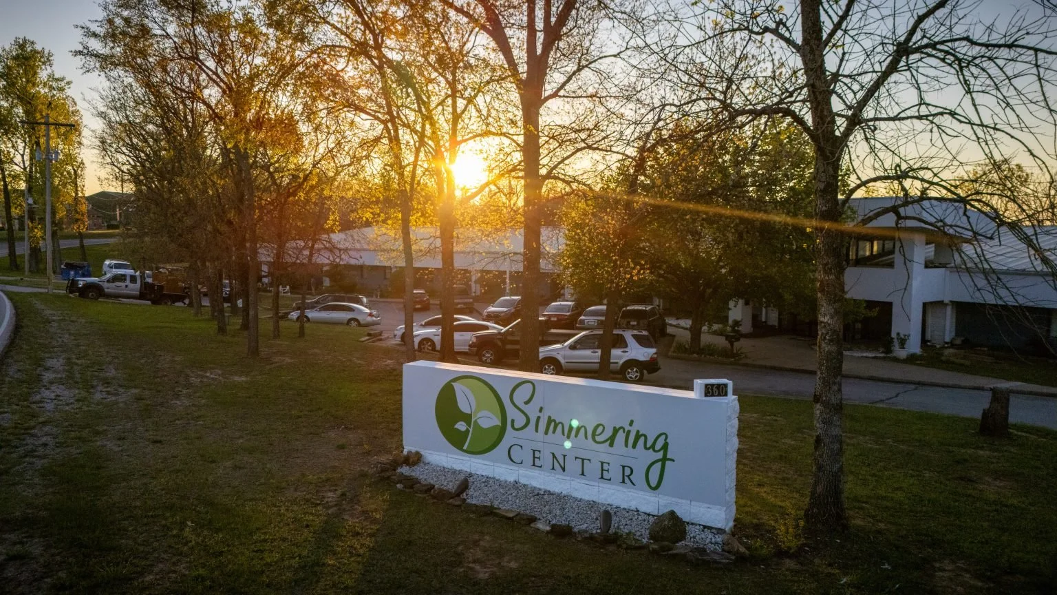 The image shows a sunset scene outside a building with a sign that reads 'Simmering Center,' featuring a green leaf logo. There are trees, parked cars, and a clear sky.