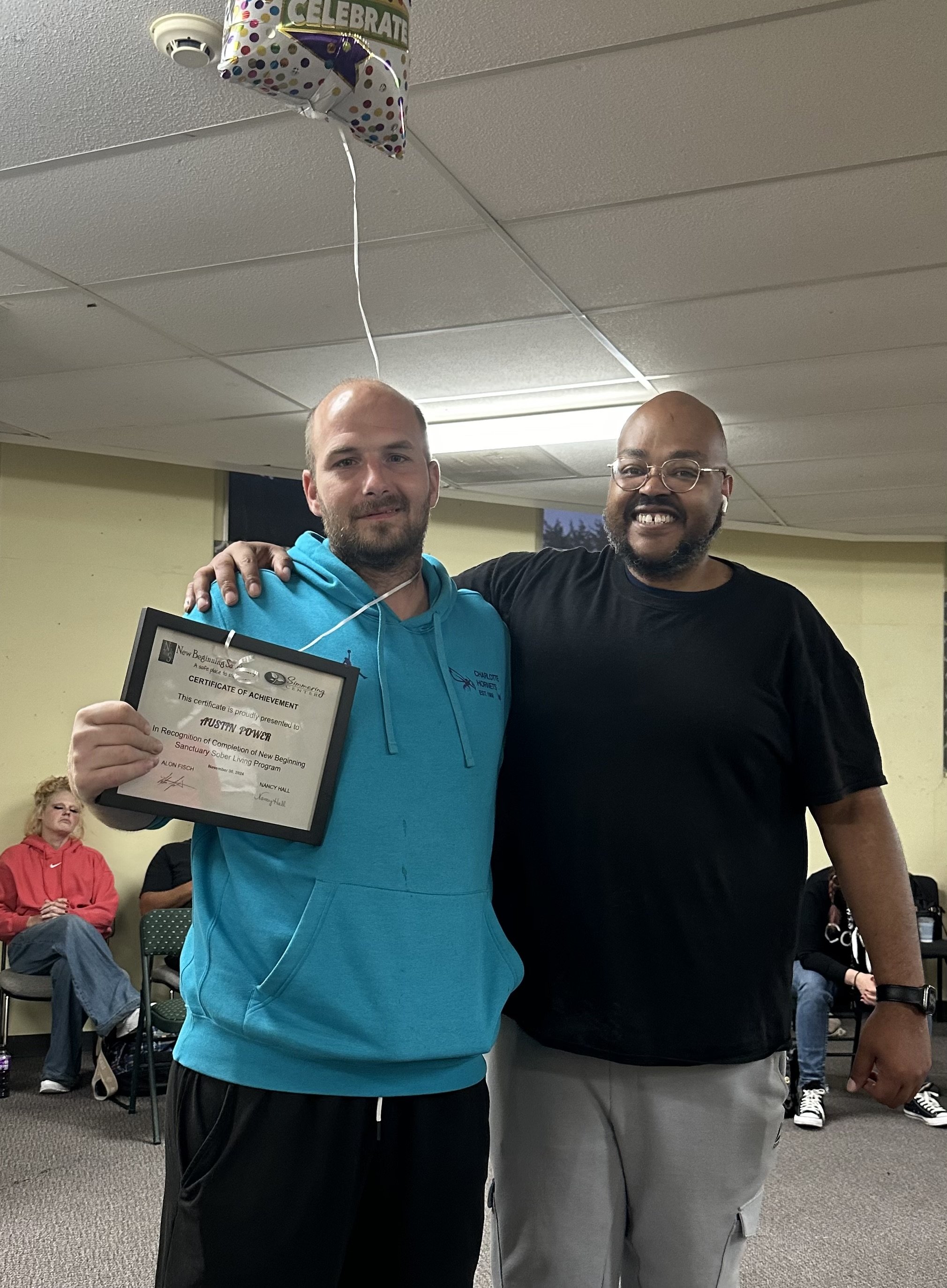 Two men standing together inside a room, smiling. The man on the left is holding a certificate and has his arm around the other man. The room has a light-colored wall, a window, and some chairs with seated people in the background. A balloon with celebratory text is floating above them.