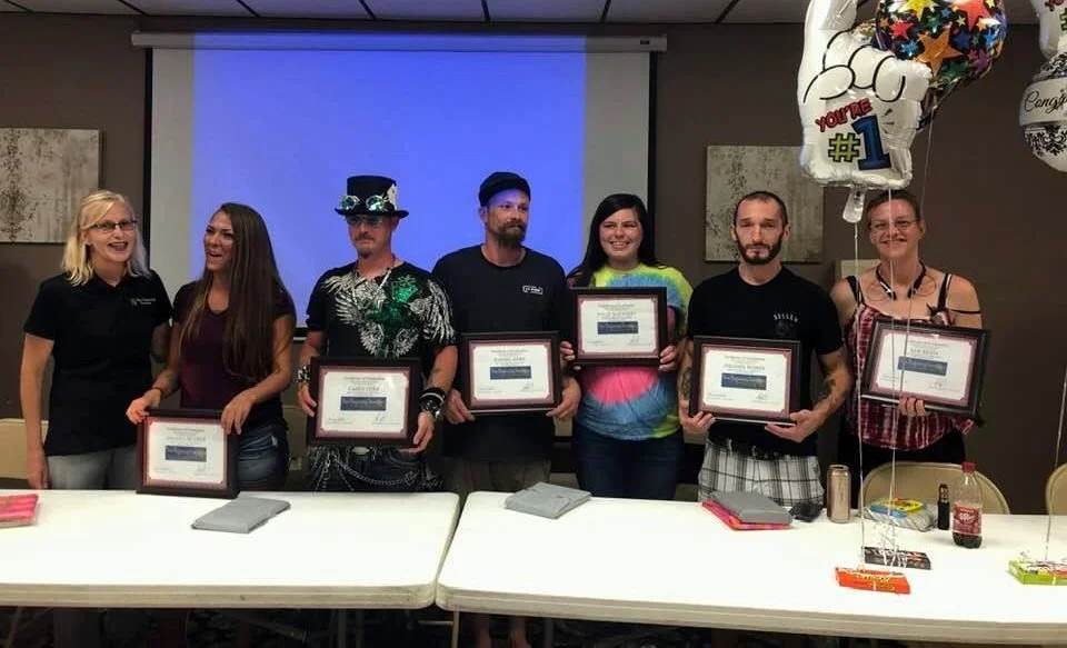 Group of six people standing behind a table, holding framed certificates, at an indoor celebration event.