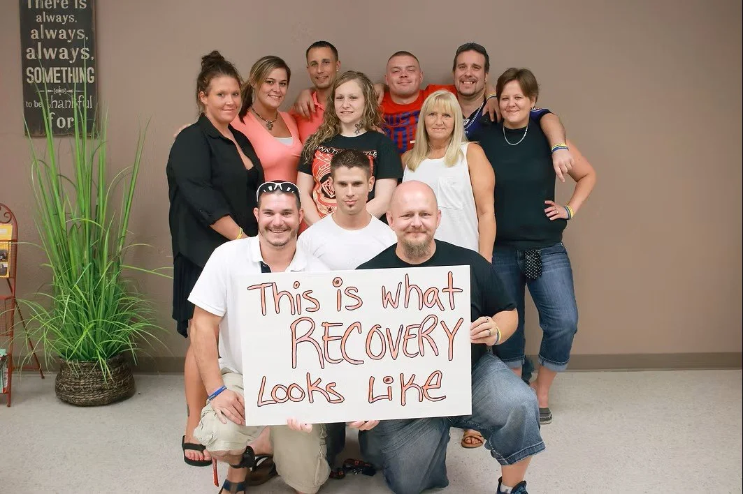 Group of ten people, smiling and standing together indoors, holding a sign that reads "This is what recovery looks like."