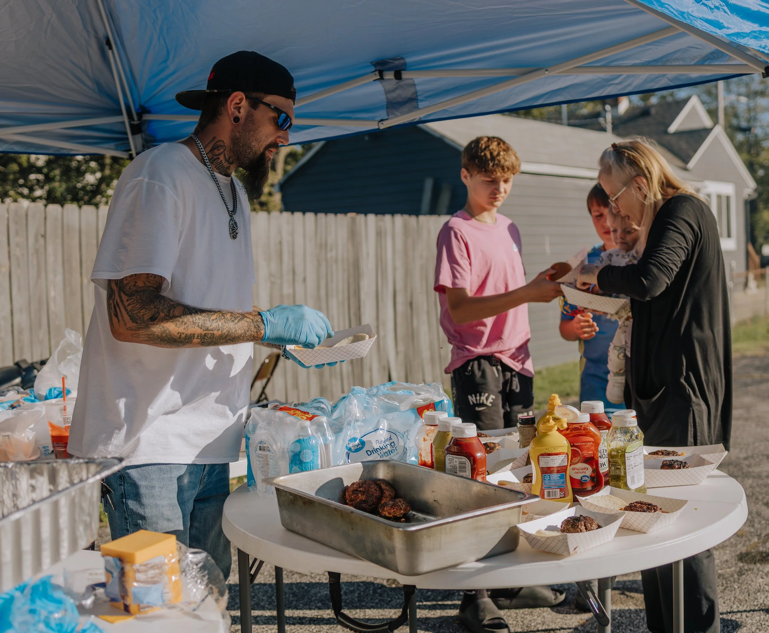 People serving and receiving food at an outdoor cookout under a blue canopy, with various condiments, water bottles, and grilled food on the table.