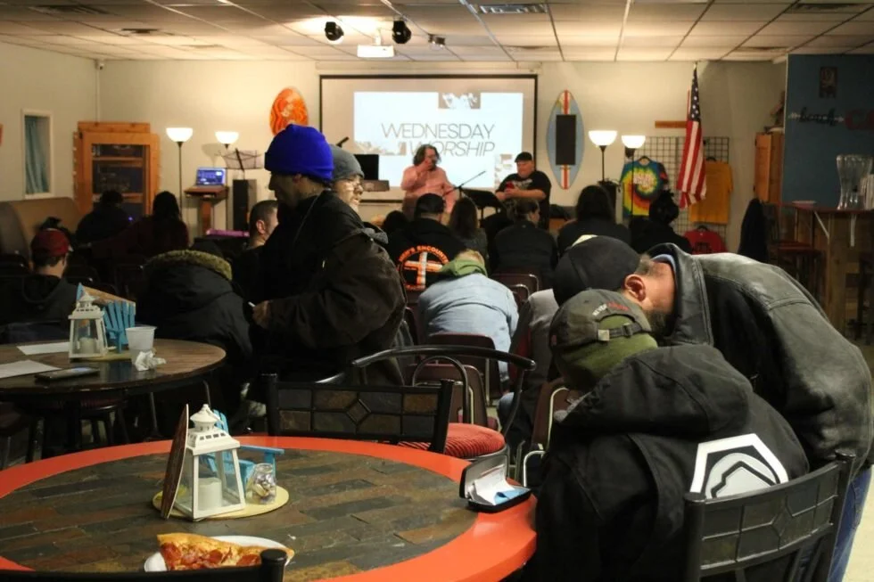 Indoor church event with people seated, some standing, listening to a speaker onstage in front of a screen that reads 'Wednesday Worship'.