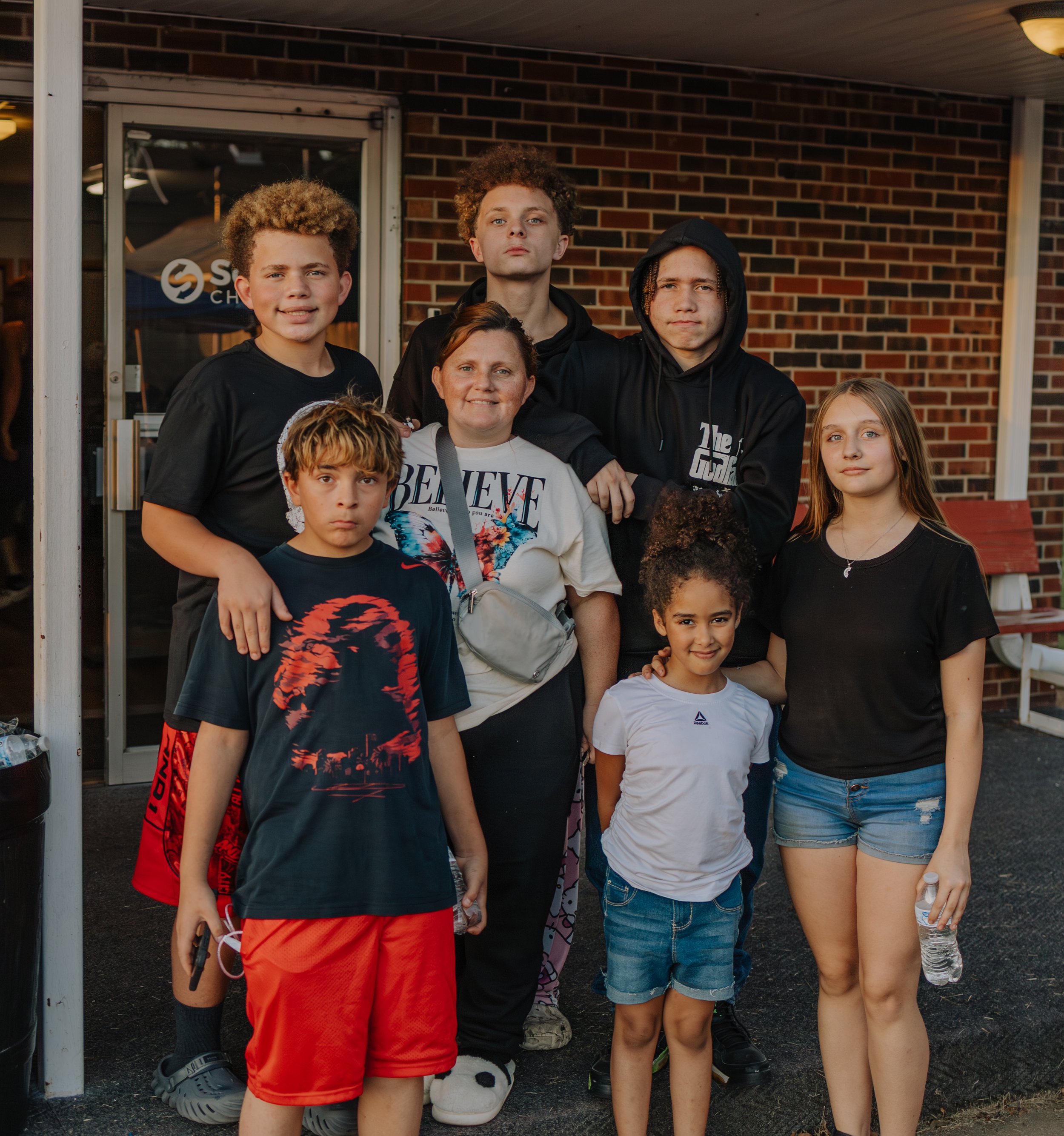 Group of nine children and one adult woman standing outside in front of a brick building, some smiling and some with neutral expressions.