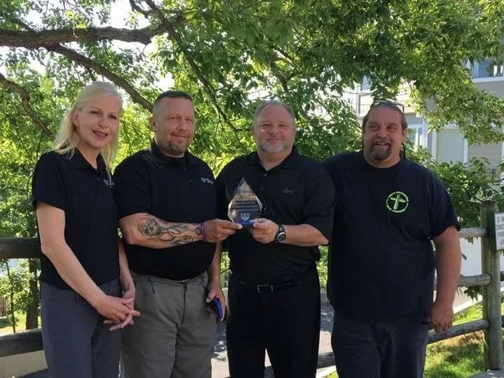 Four people standing outdoors under a tree, two men are holding a glass award, the group is smiling and dressed in dark casual clothing.