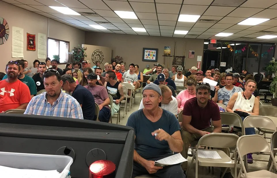A community meeting with around 50 people seated in a room, some taking notes, listening attentively, with decorations and windows in the background.