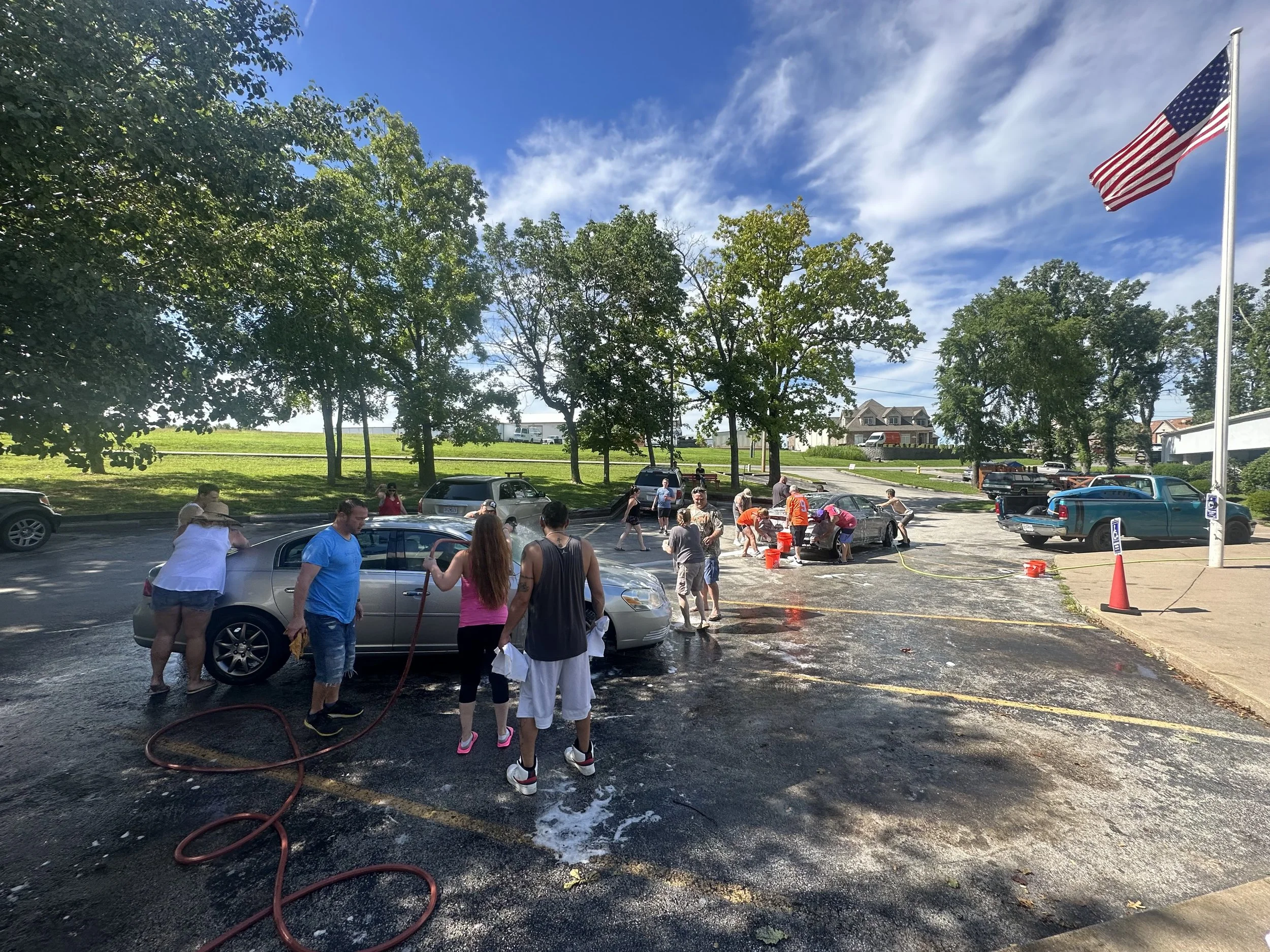 People washing cars in a parking lot on a sunny day, with trees, a grassy area, and an American flag in the background.