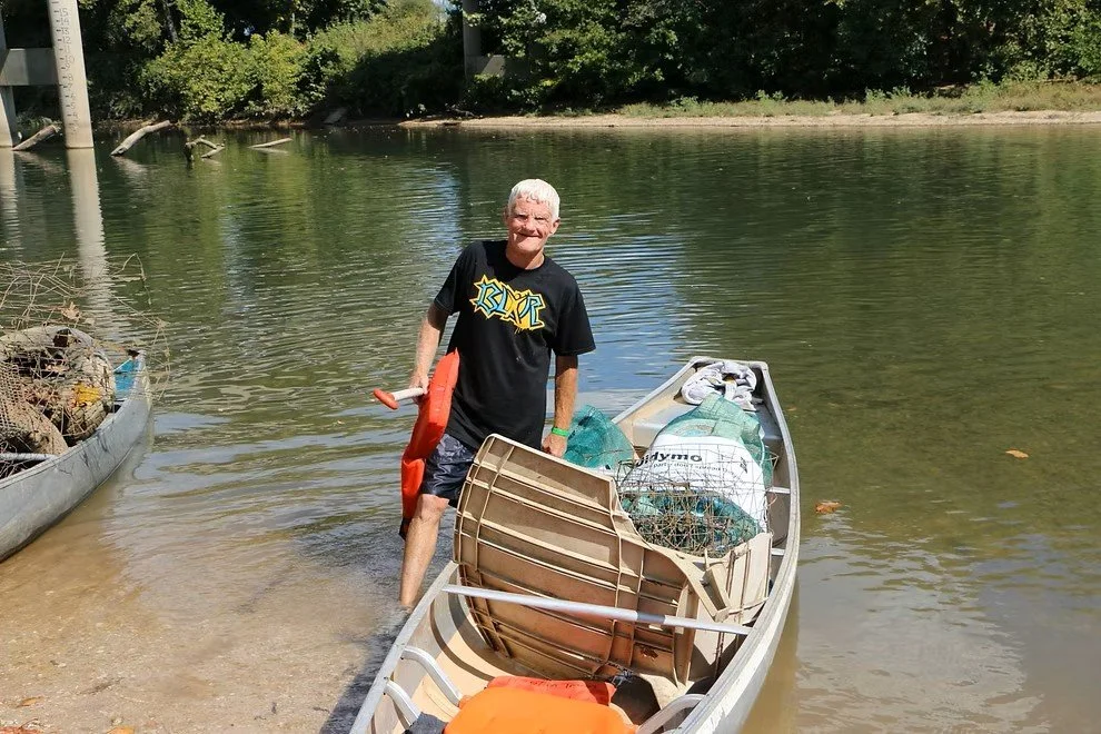A man with gray hair and a black T-shirt standing in shallow water next to a boat loaded with supplies and gear, near a wooded shoreline.