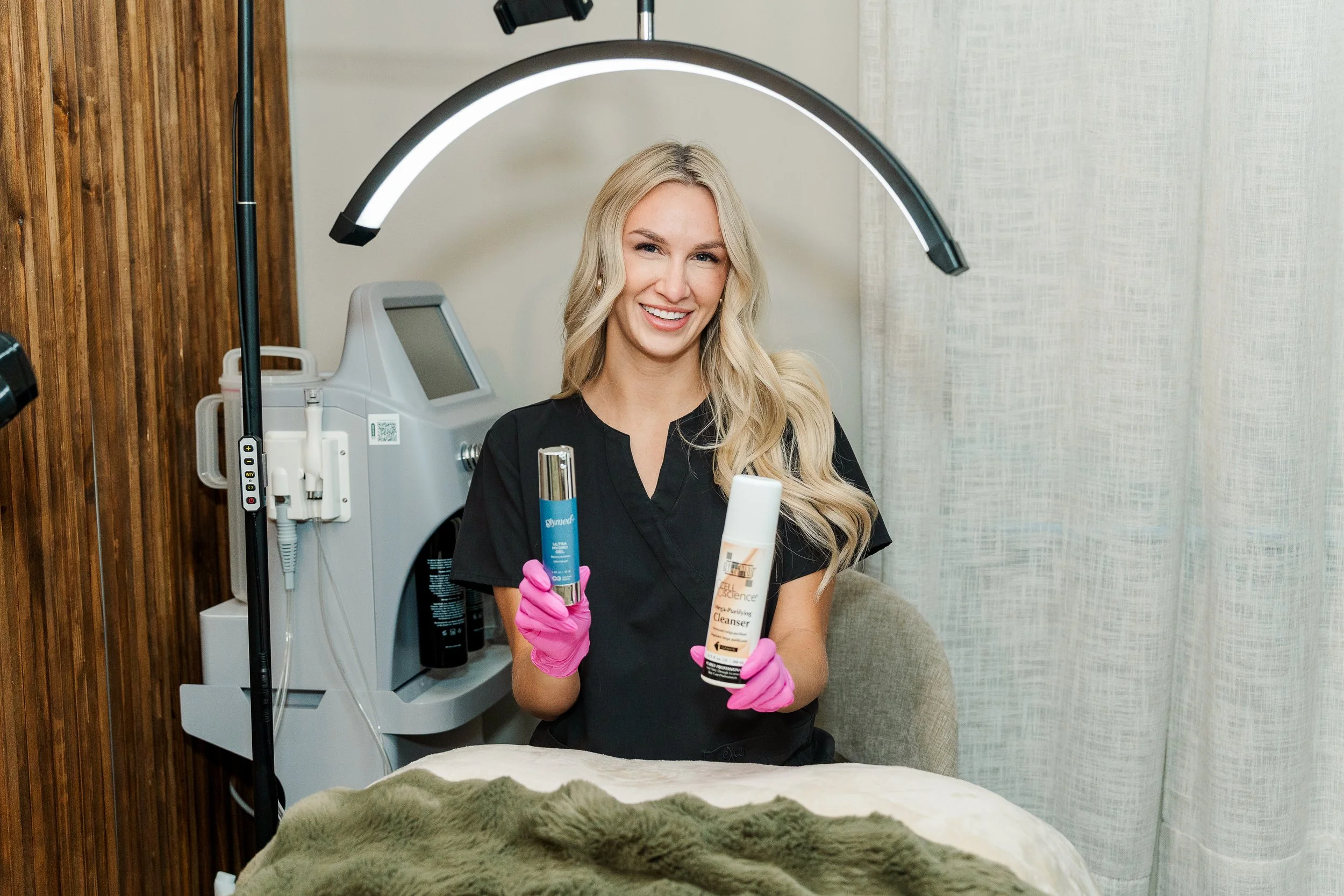 A smiling woman in black scrubs wearing pink gloves holding skincare products in a clinic or spa setting.