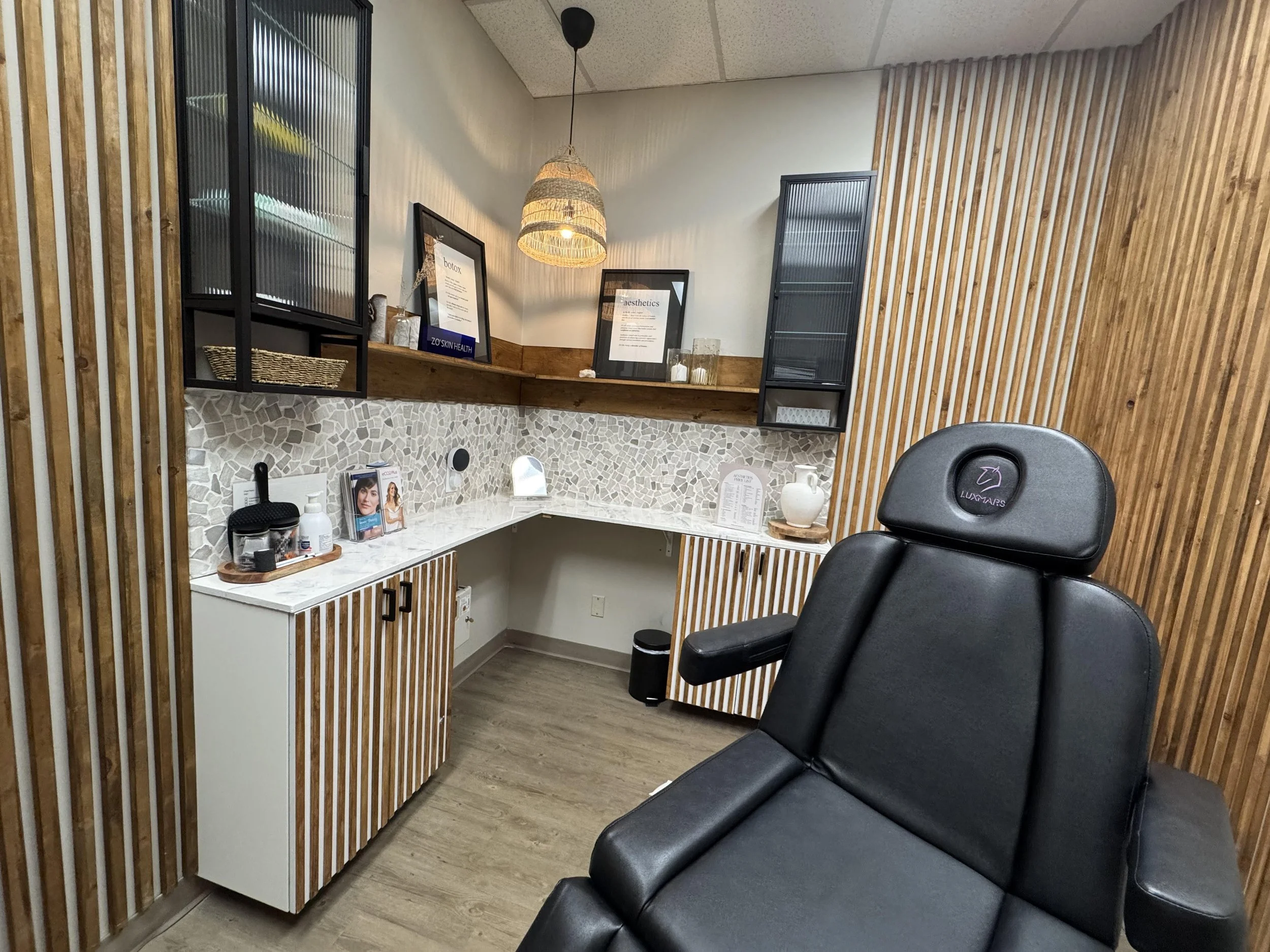 A cozy spa treatment room with a black massage chair, wooden slat walls, and a marble-topped cabinet with beauty products and informational brochures.