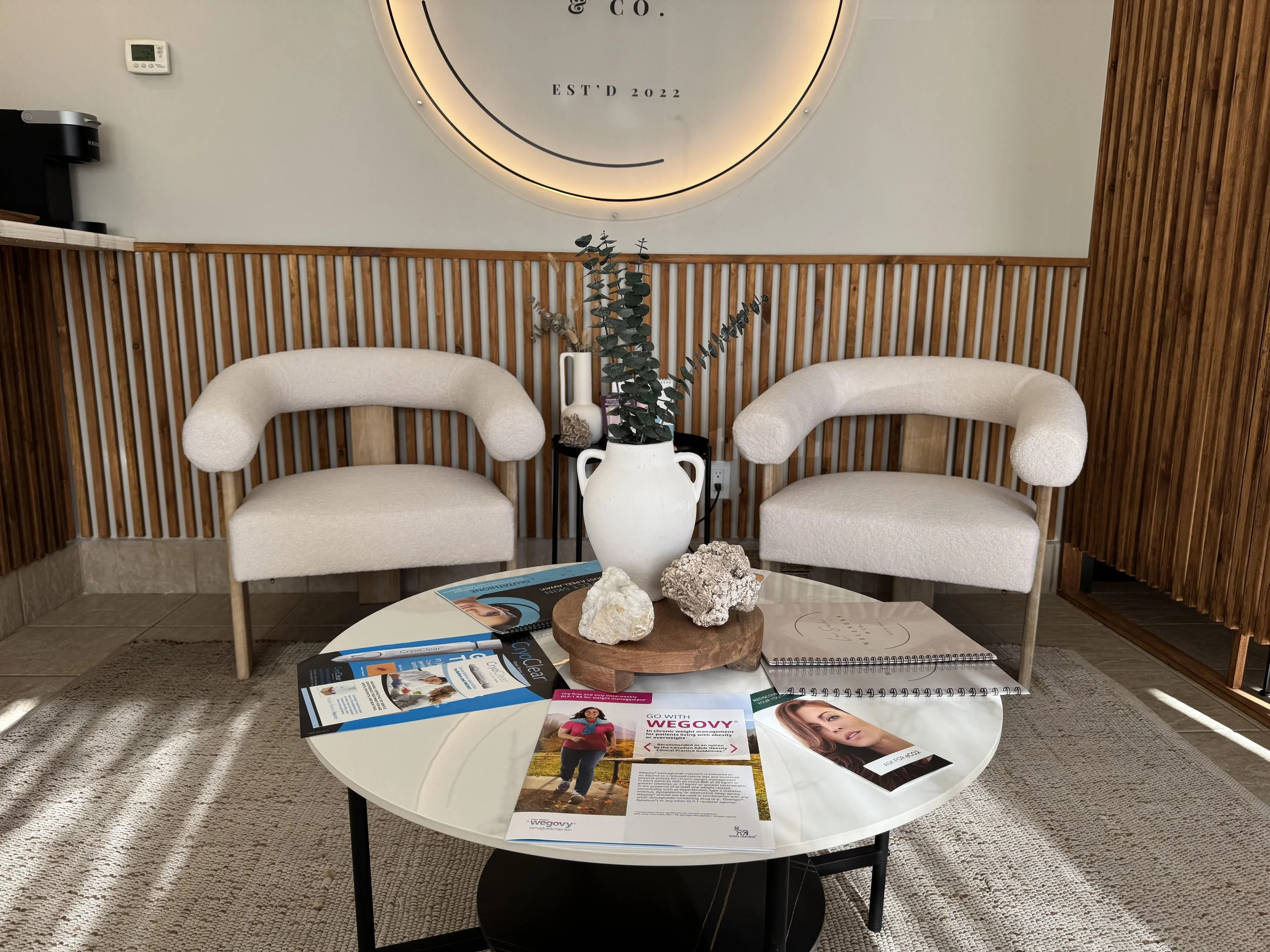 A seating area with two cream-colored chairs with curved arms facing each other, a round white coffee table in the center, and a wooden wall with a circular sign behind. The table holds a large white vase with green foliage, rocks, brochures, and a n