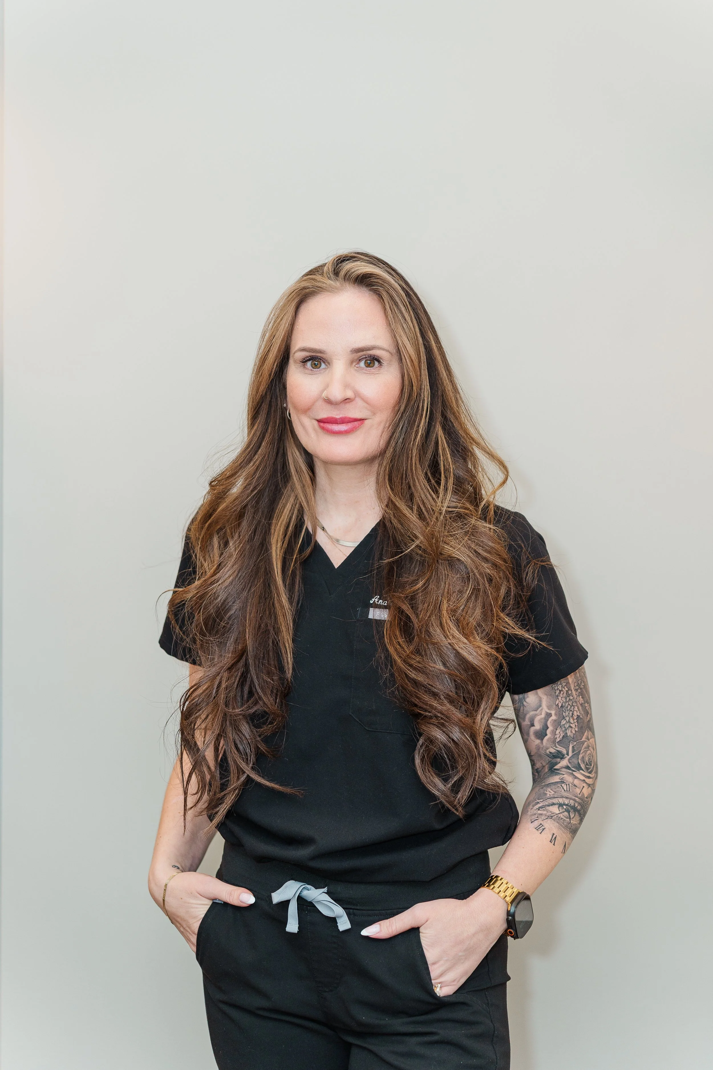 A woman with long, wavy brown hair, wearing a black medical uniform, standing against a plain light-colored wall, looking at the camera with a slight smile.