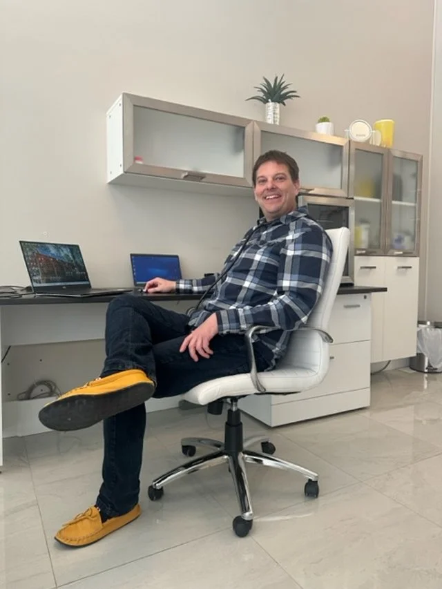 A man sitting in a white office chair at a desk with two laptops, smiling at the camera, in a room with white and gray furniture and a potted plant on a shelf.