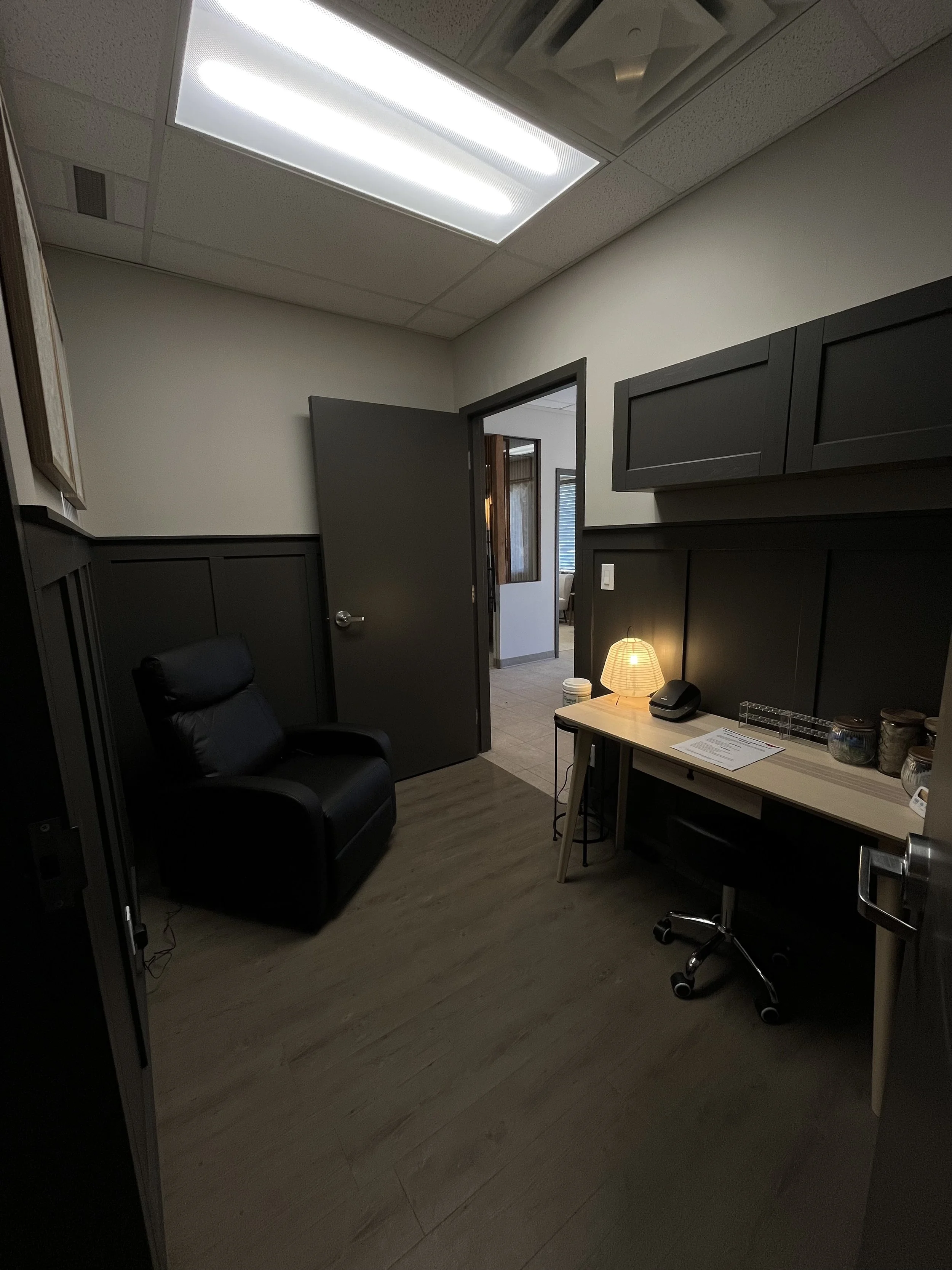 An office room with a black reclining chair, a small desk with a chair, a lamp, and some jars, with a door leading to a hallway or another room. The office has dark gray wainscoting and cabinetry, a light-colored wall, and wood flooring.