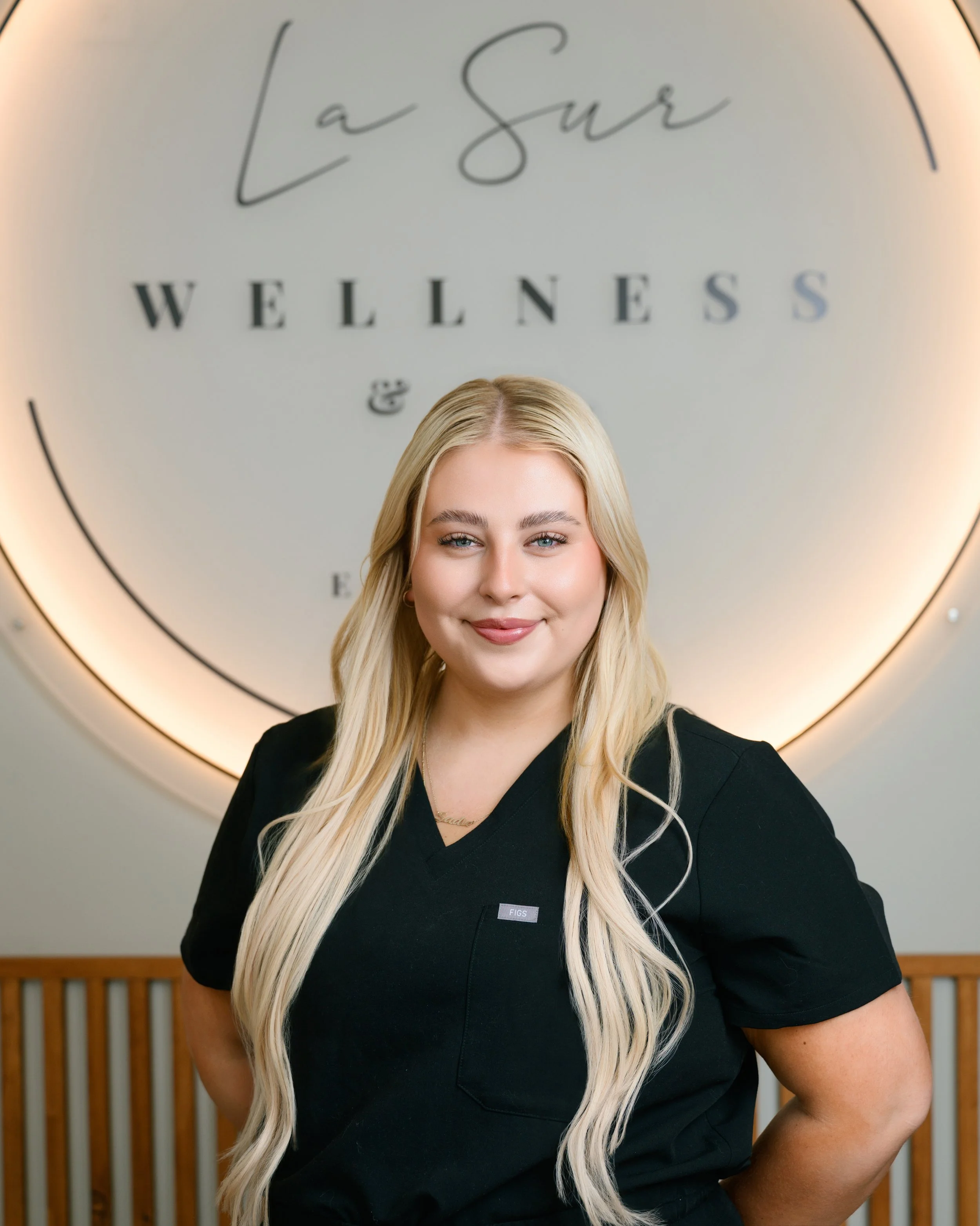 Portrait of a young blonde woman smiling, wearing a black shirt and a necklace, standing against a plain white background.