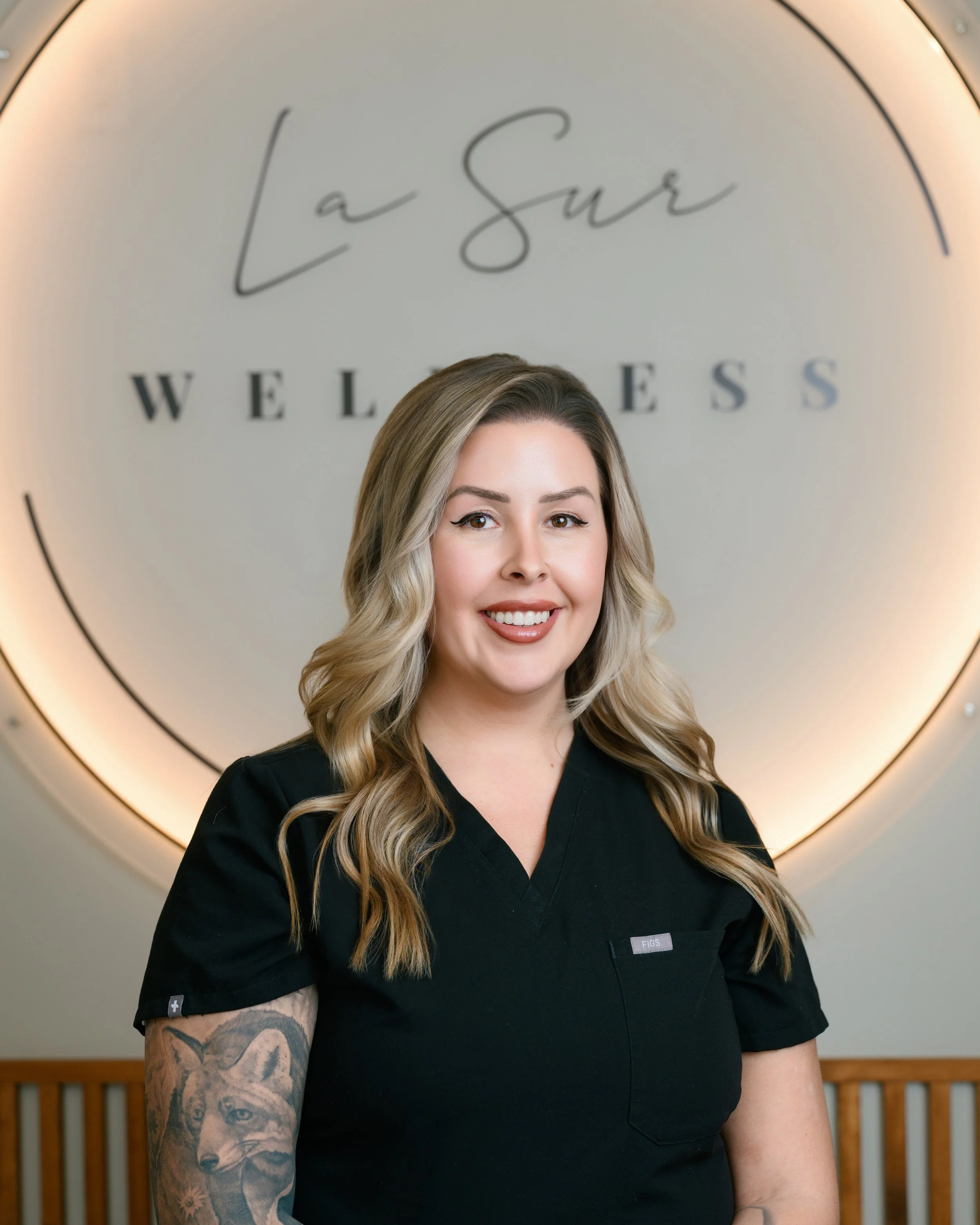 Female healthcare professional with tattoos, arms crossed, wearing a black medical scrub top, standing against a plain light-colored wall.