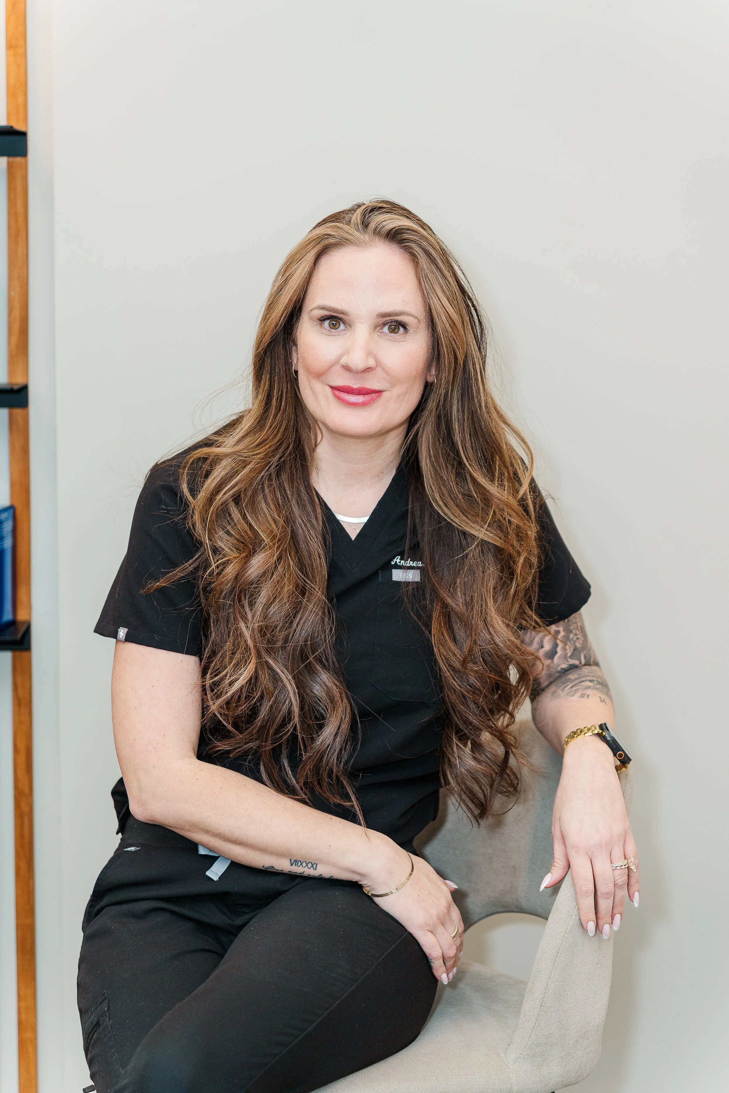 A woman with long wavy brown hair sitting on a beige chair in a medical uniform, smiling at the camera.