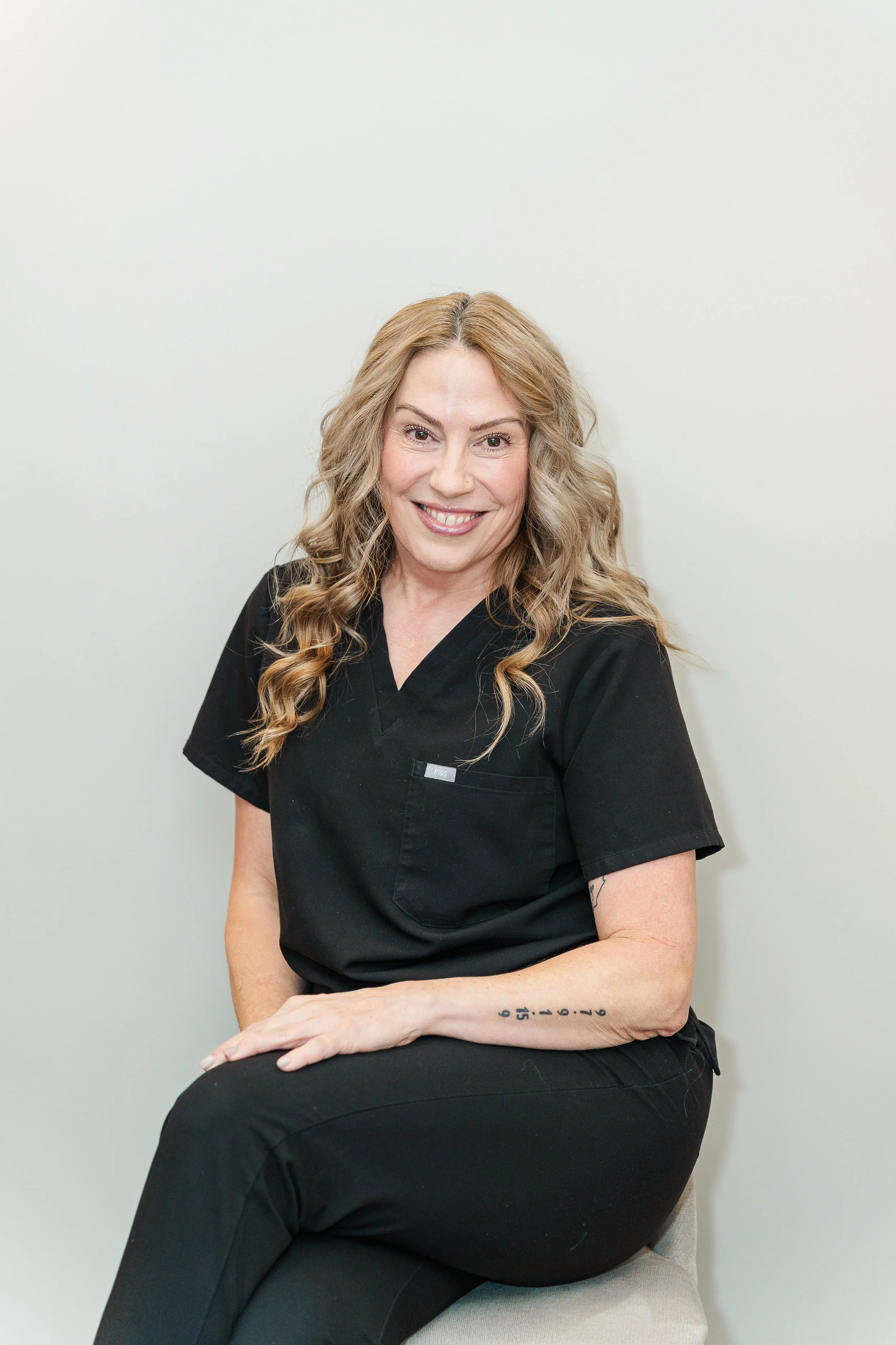 A woman with long, wavy blonde hair, wearing a black medical uniform, sitting on a beige stool against a plain off-white wall, smiling at the camera.
