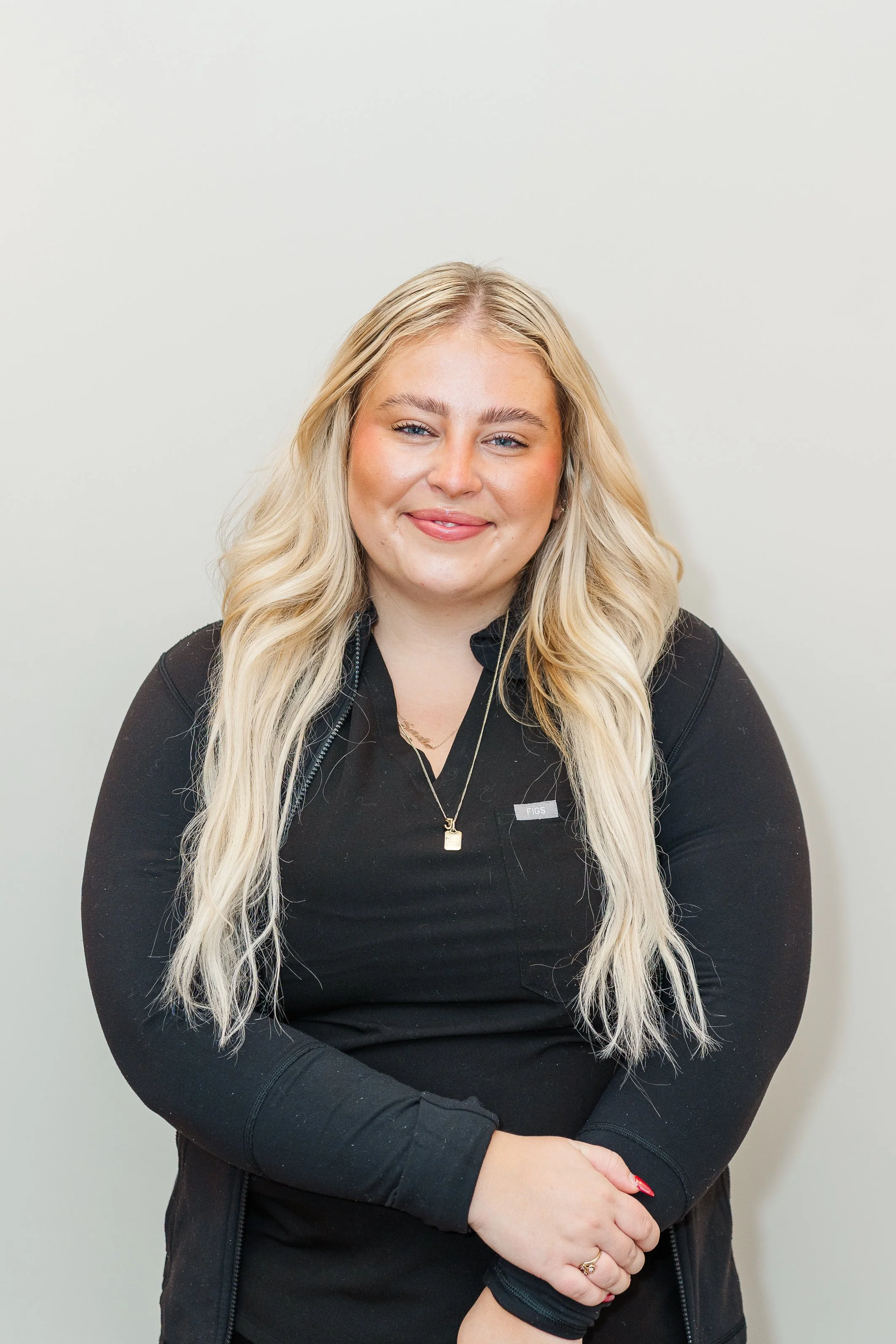 Portrait of a young blonde woman smiling, wearing a black shirt and a necklace, standing against a plain white background.