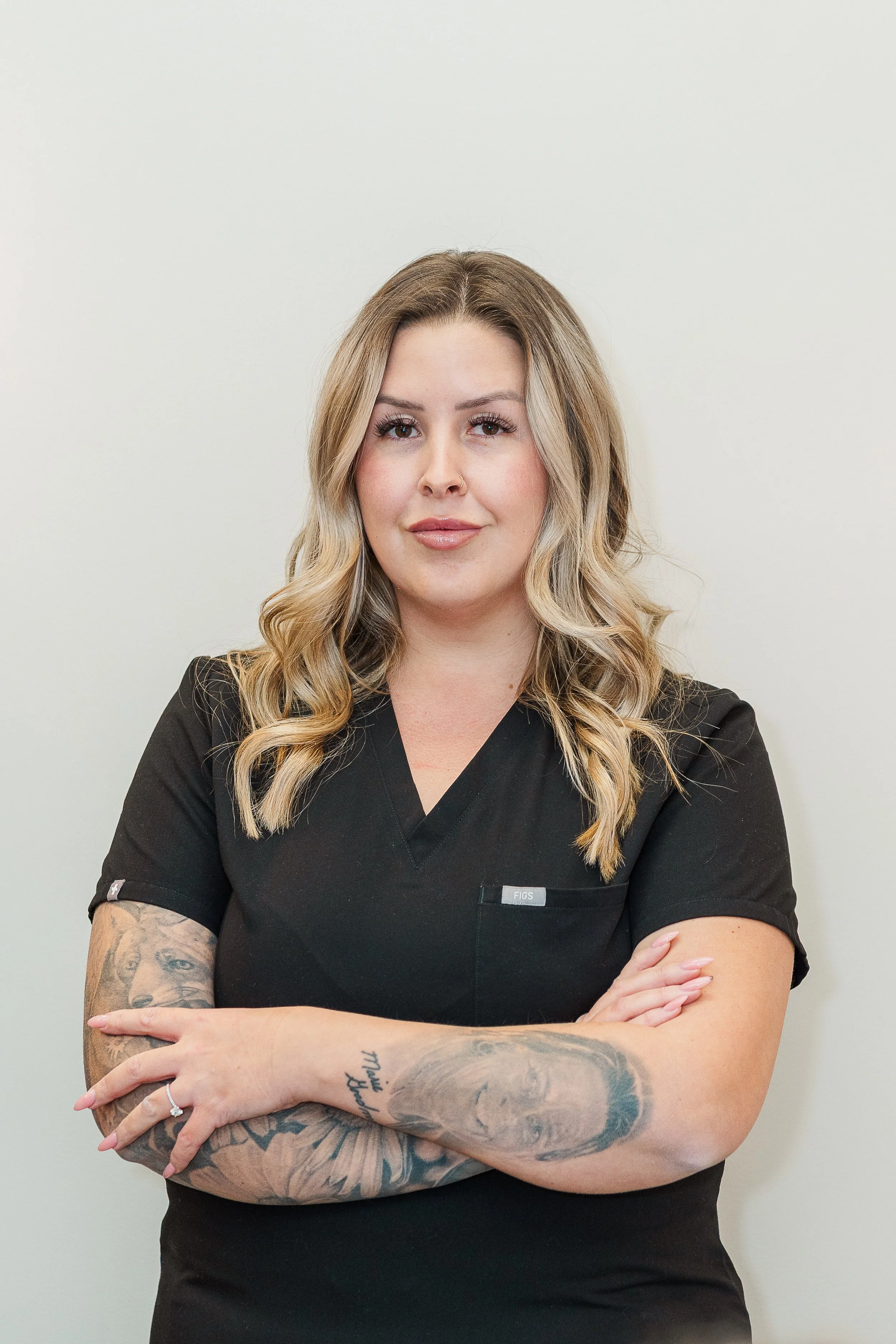 Female healthcare professional with tattoos, arms crossed, wearing a black medical scrub top, standing against a plain light-colored wall.
