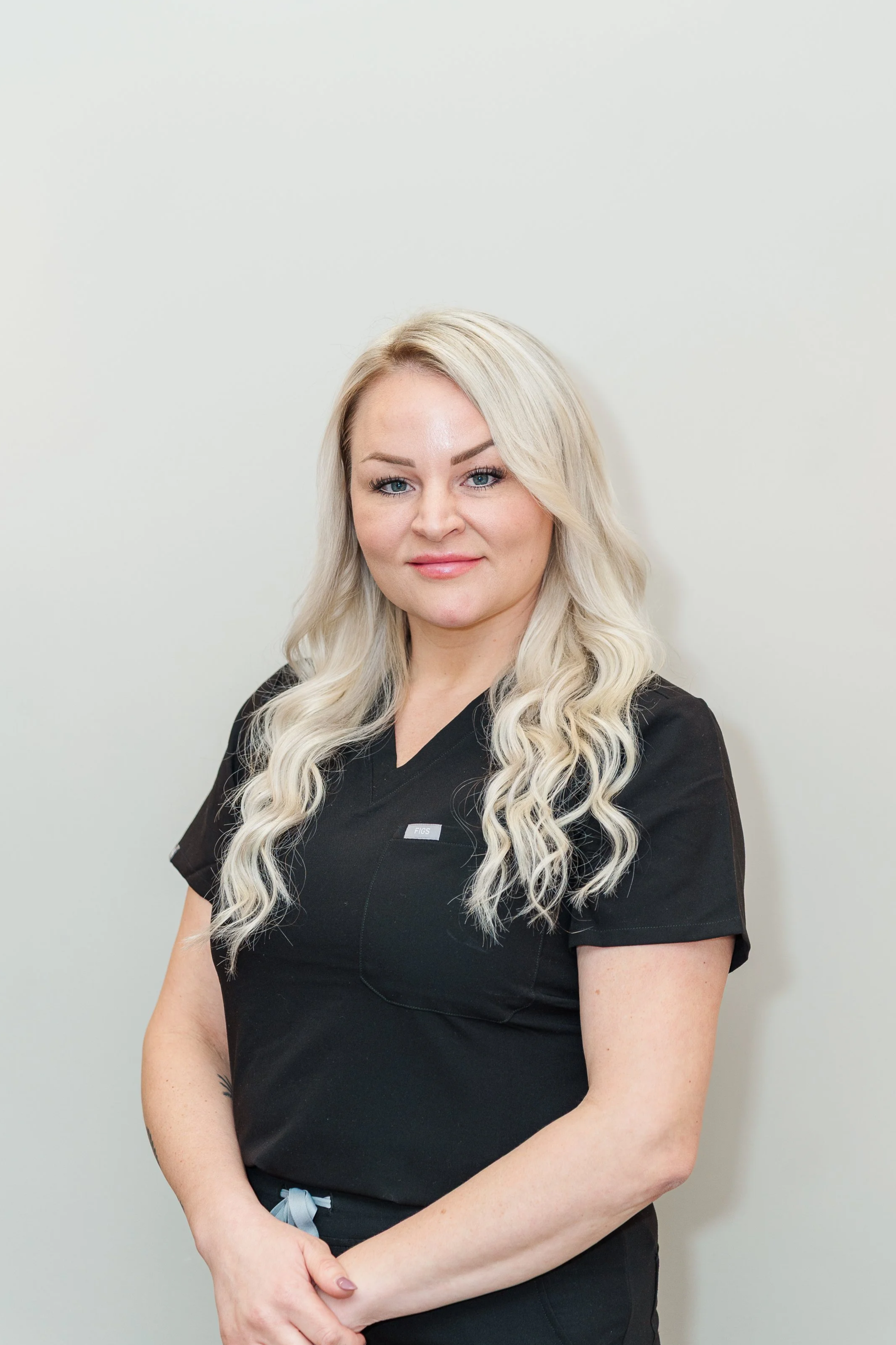 A woman with long, wavy platinum blonde hair wearing black medical scrubs standing against a plain white wall.