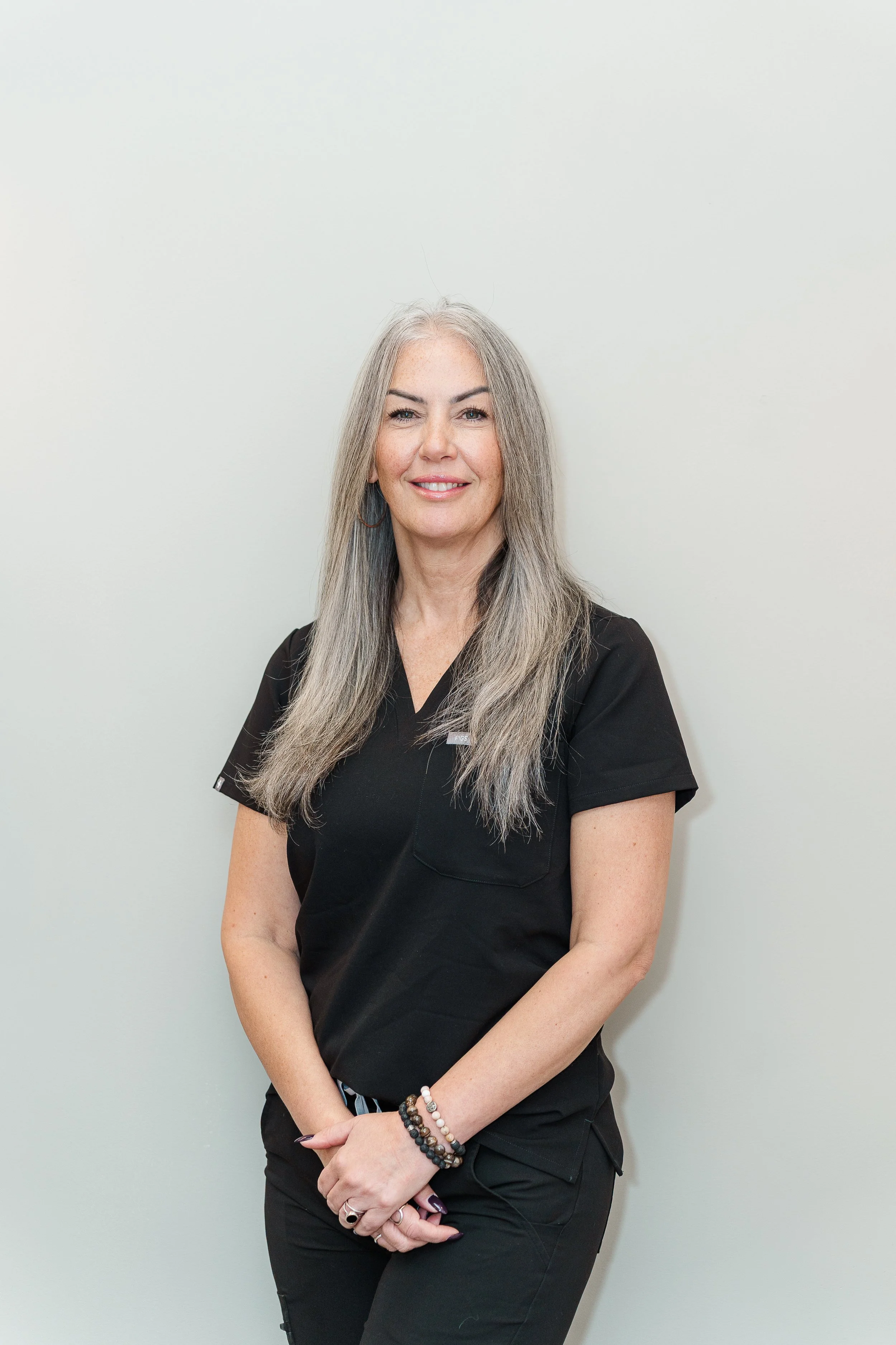 A middle-aged woman with long gray hair and light skin, wearing black scrubs and looking confidently at the camera, standing against a plain light-colored wall.