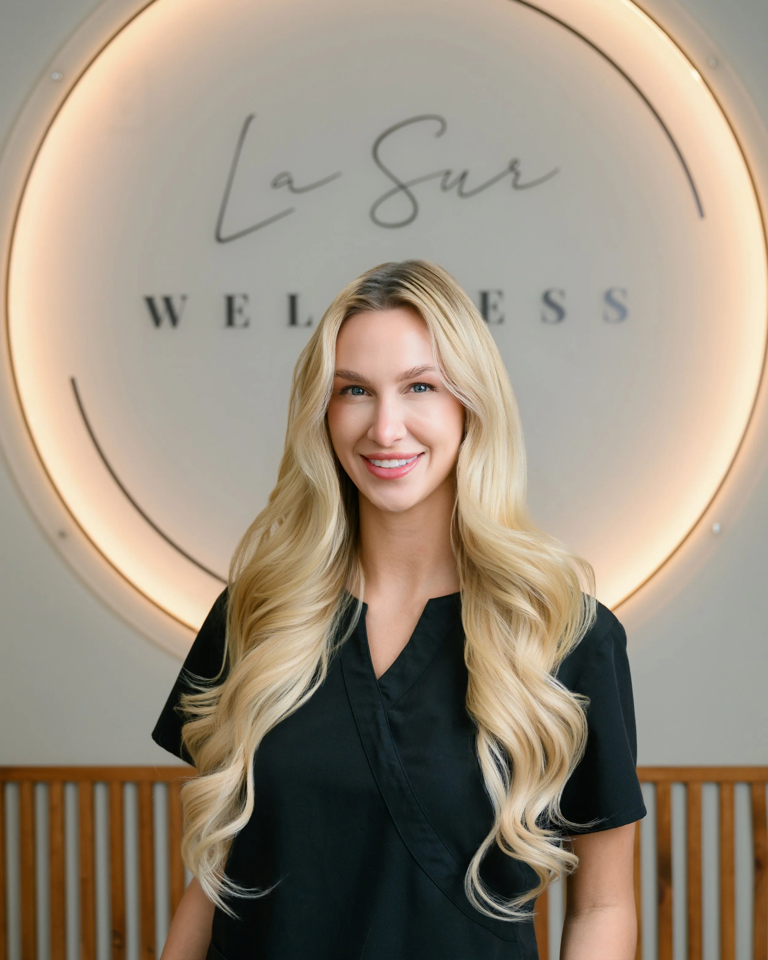 A woman with long blonde hair, smiling, wearing black scrubs, sitting on a gray chair against a plain white wall.