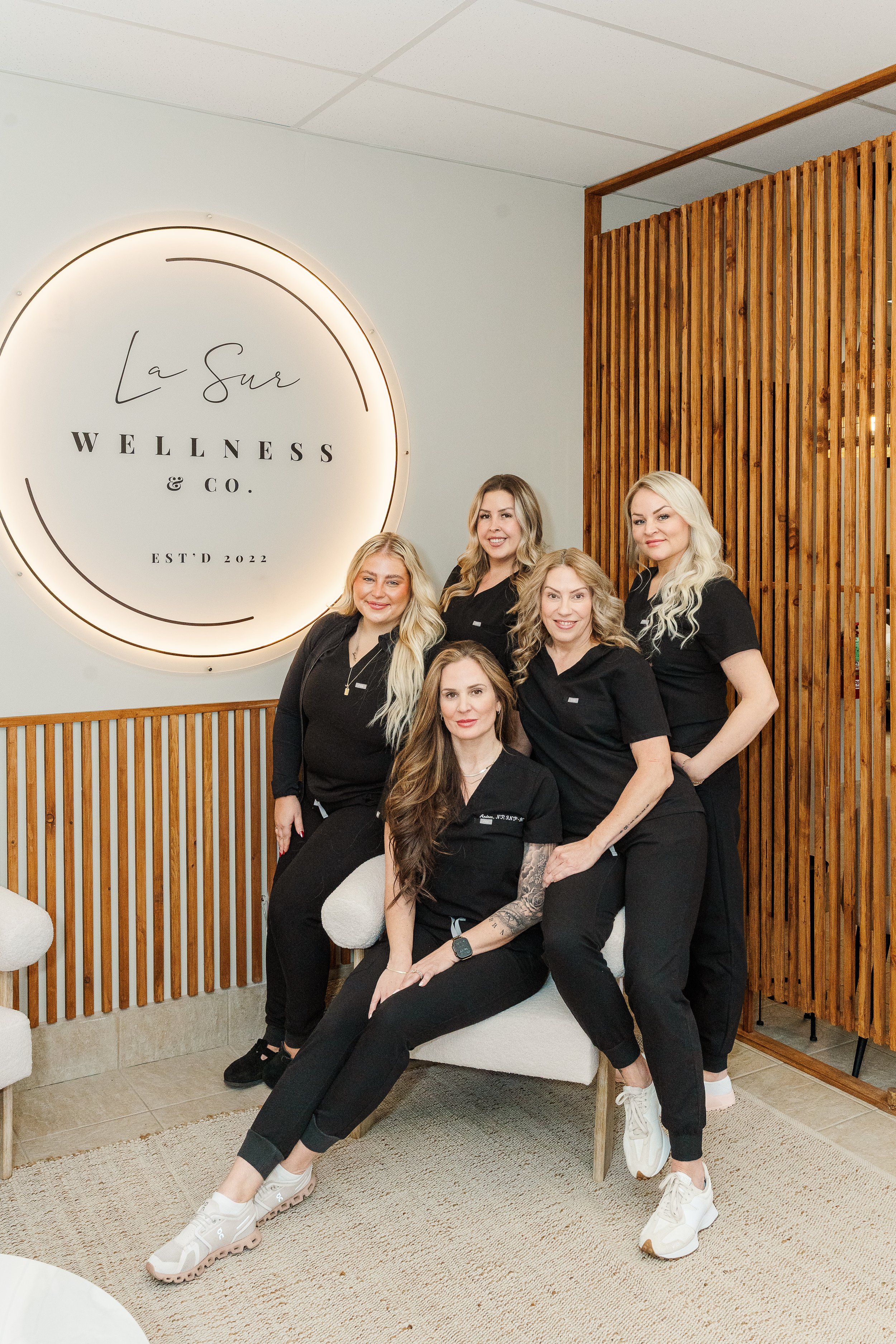 Group of five women in black uniforms sitting and standing in wellness center, with wooden wall and illuminated sign that reads 'La Sur Wellness & Co.'