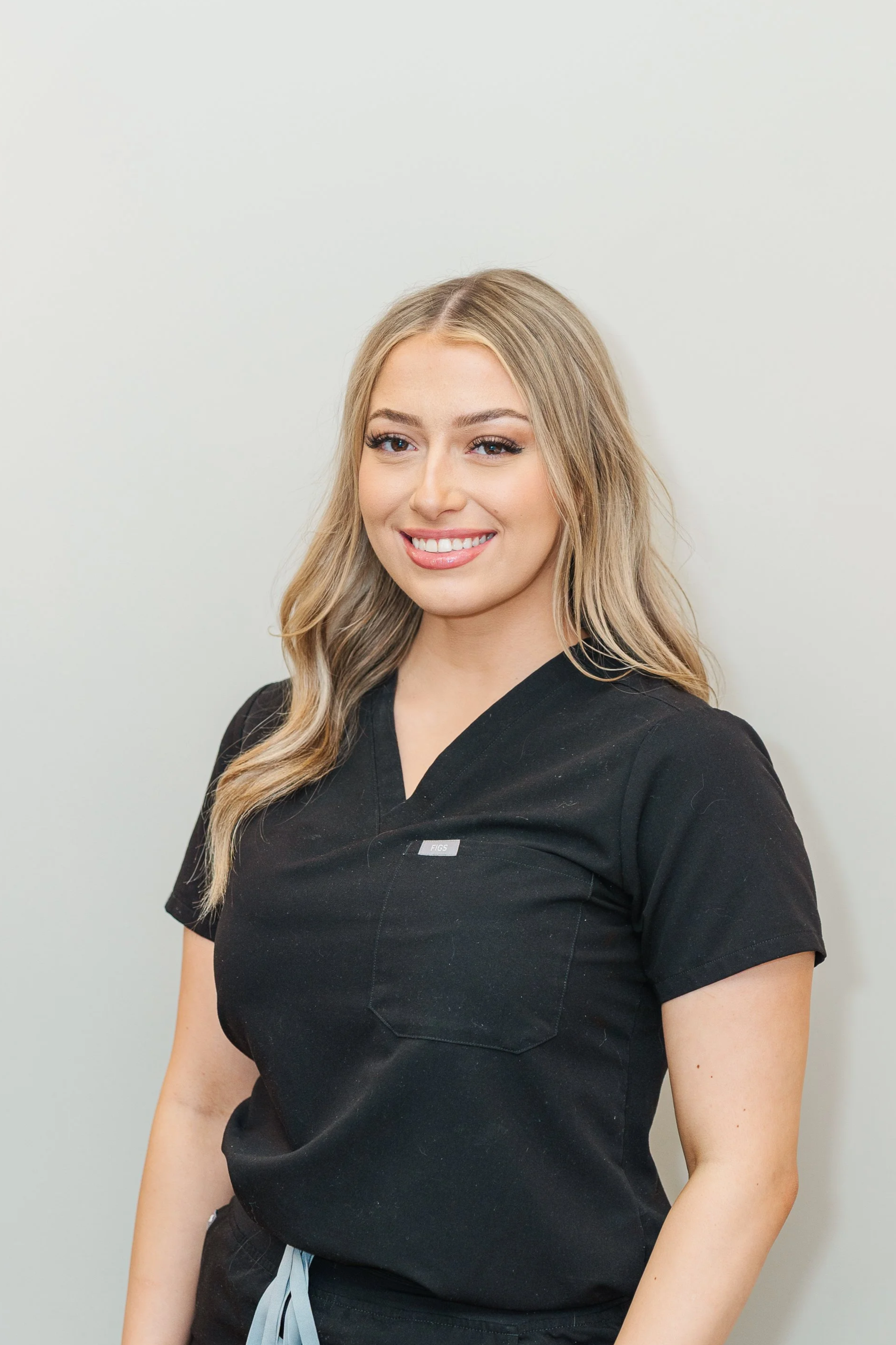 A smiling young woman with long blonde hair wearing black scrubs, standing against a plain white wall.