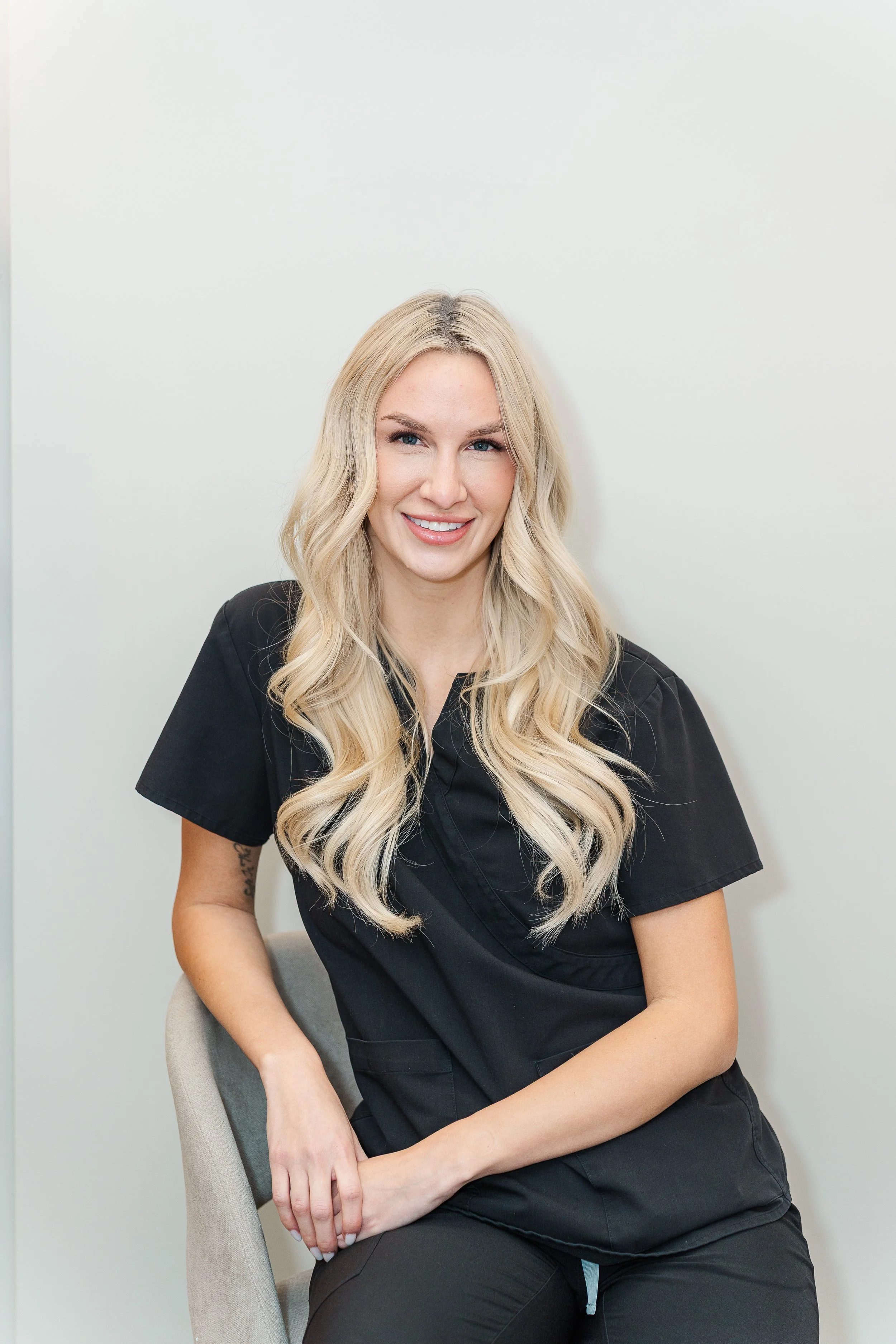 A woman with long blonde hair, smiling, wearing black scrubs, sitting on a gray chair against a plain white wall.