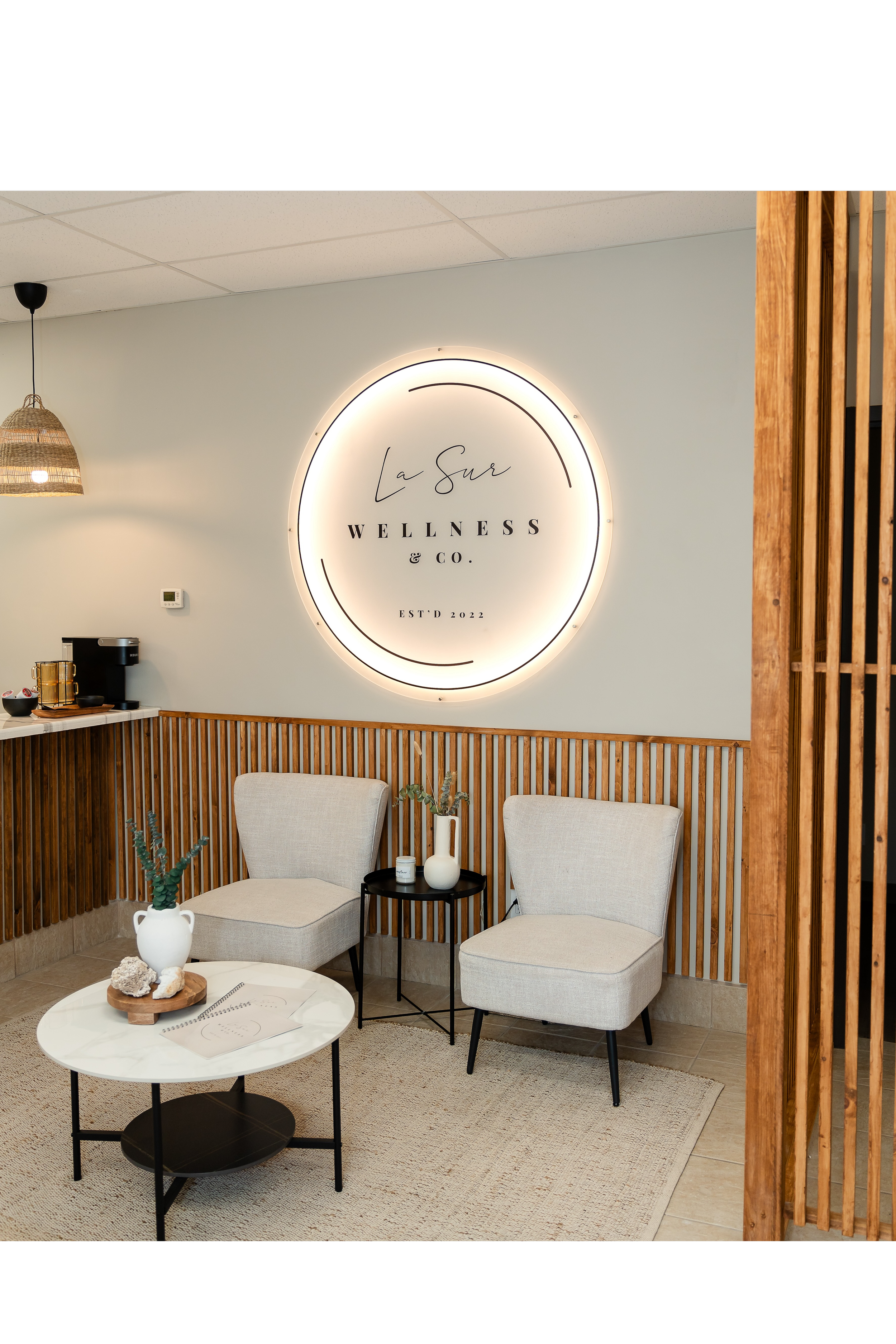 Interior of Wellness & Co. with two beige armchairs, a small black side table with vases and plants, a white marble coffee table with decorative rocks and notebooks, and a large circular sign with the business name on the wall.