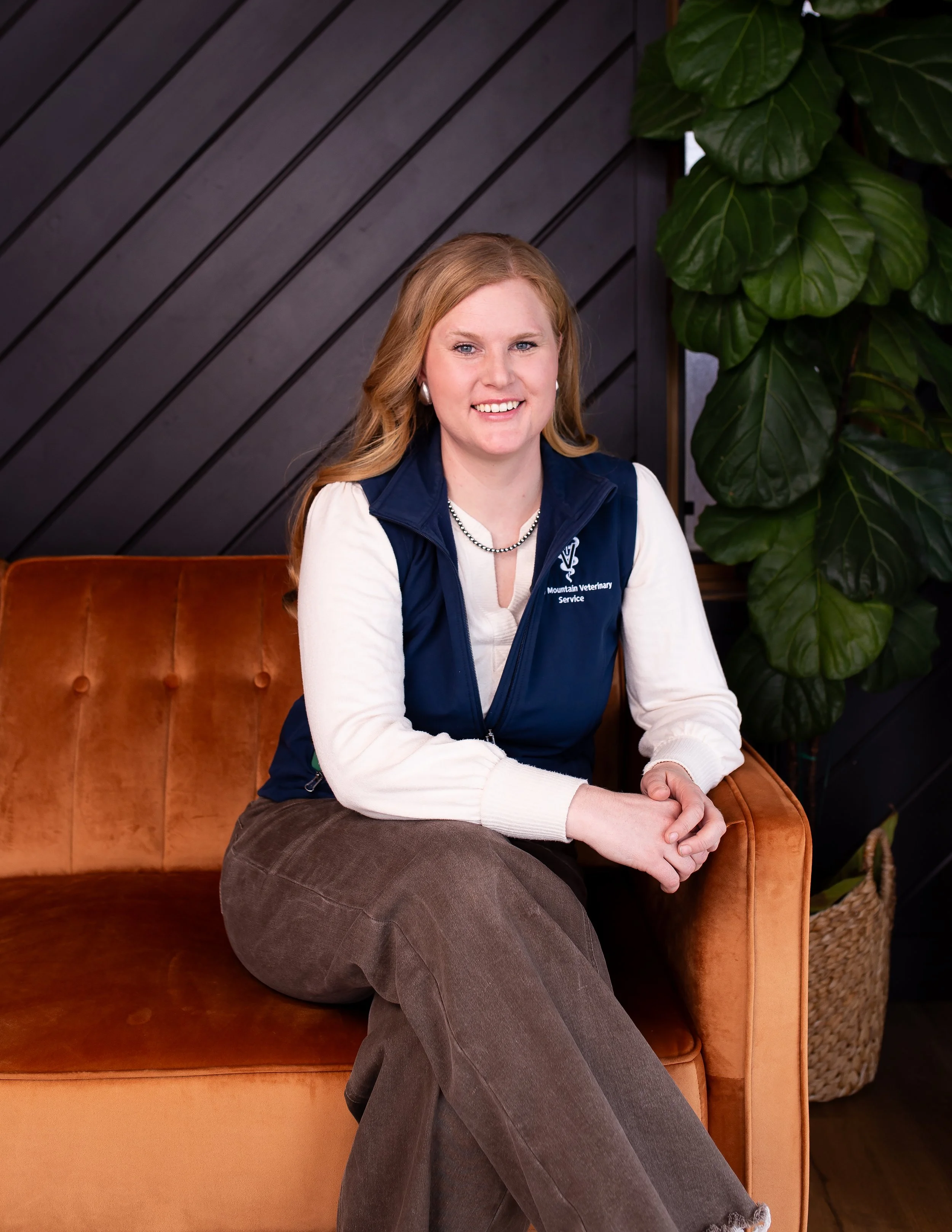 A family of five sitting on the grass in a field, with mountains and a cloudy sky in the background. The father is wearing a cowboy hat and has a beard. The mother has long red hair. Two young children, a boy and a girl, are sitting with them, smiling.