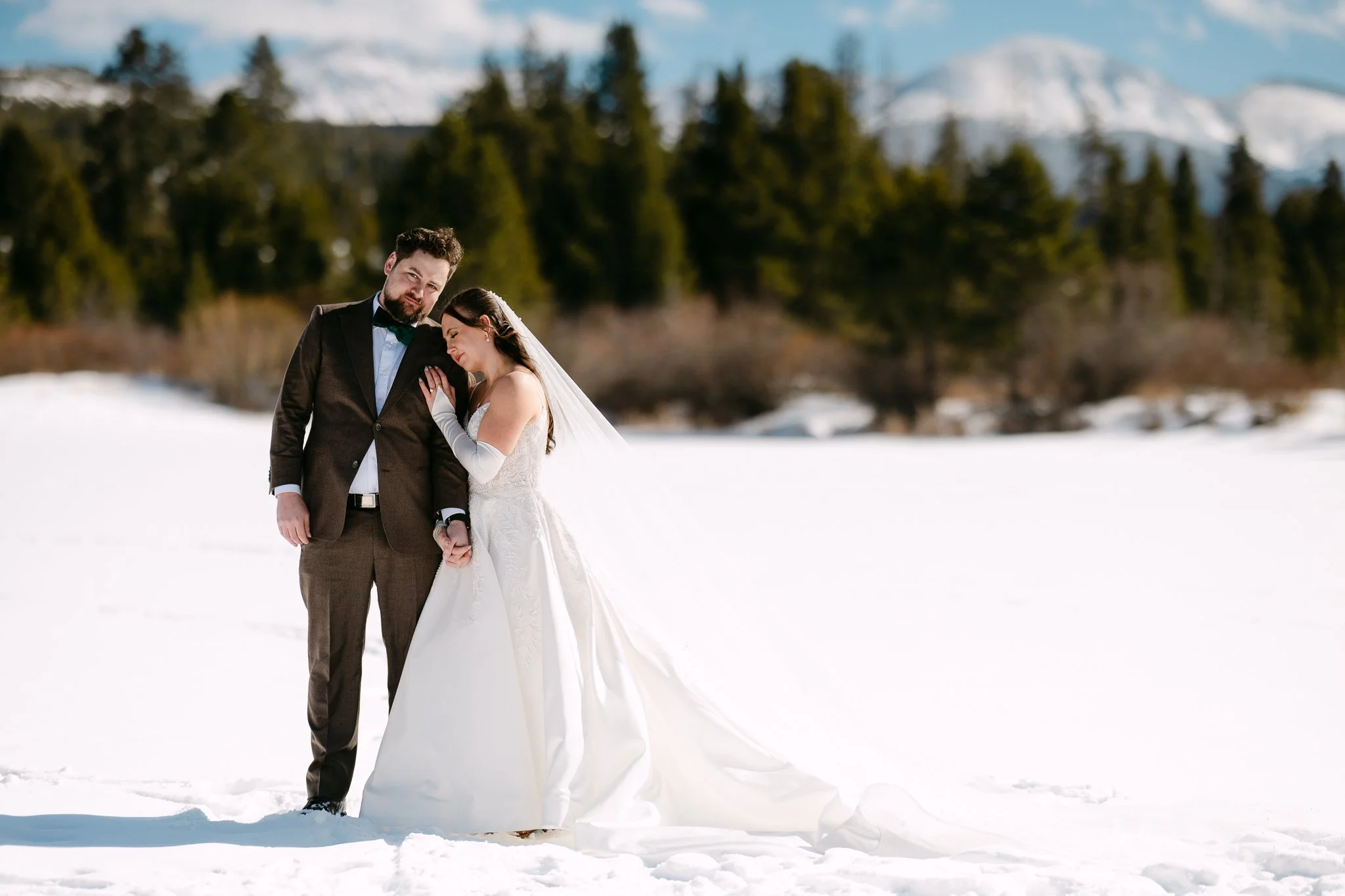 A bride and groom standing together in a snowy landscape with mountains and trees in the background, holding hands and embracing.