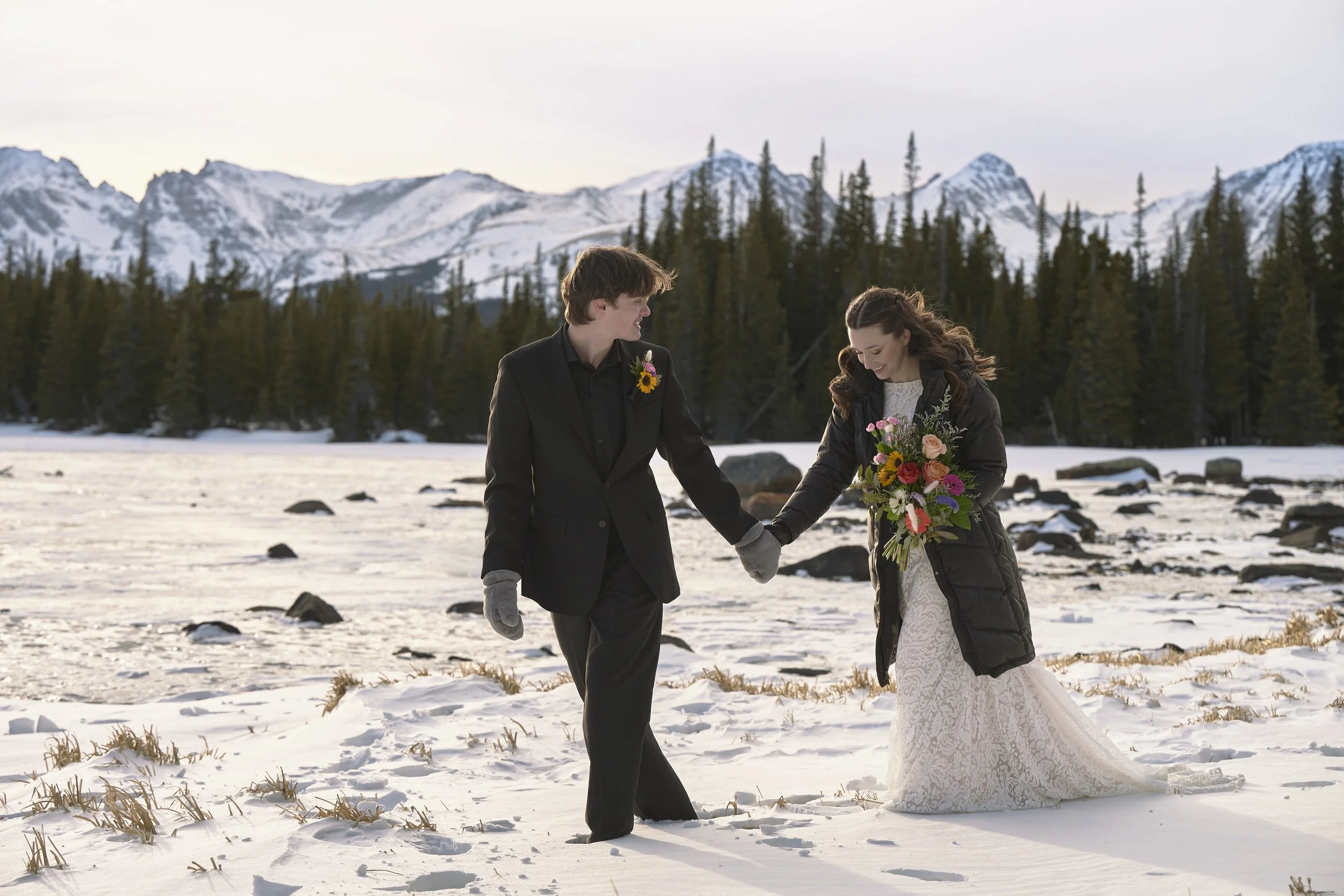 A couple dressed in wedding attire holding hands in a snowy, mountainous landscape with pine trees and a frozen river.