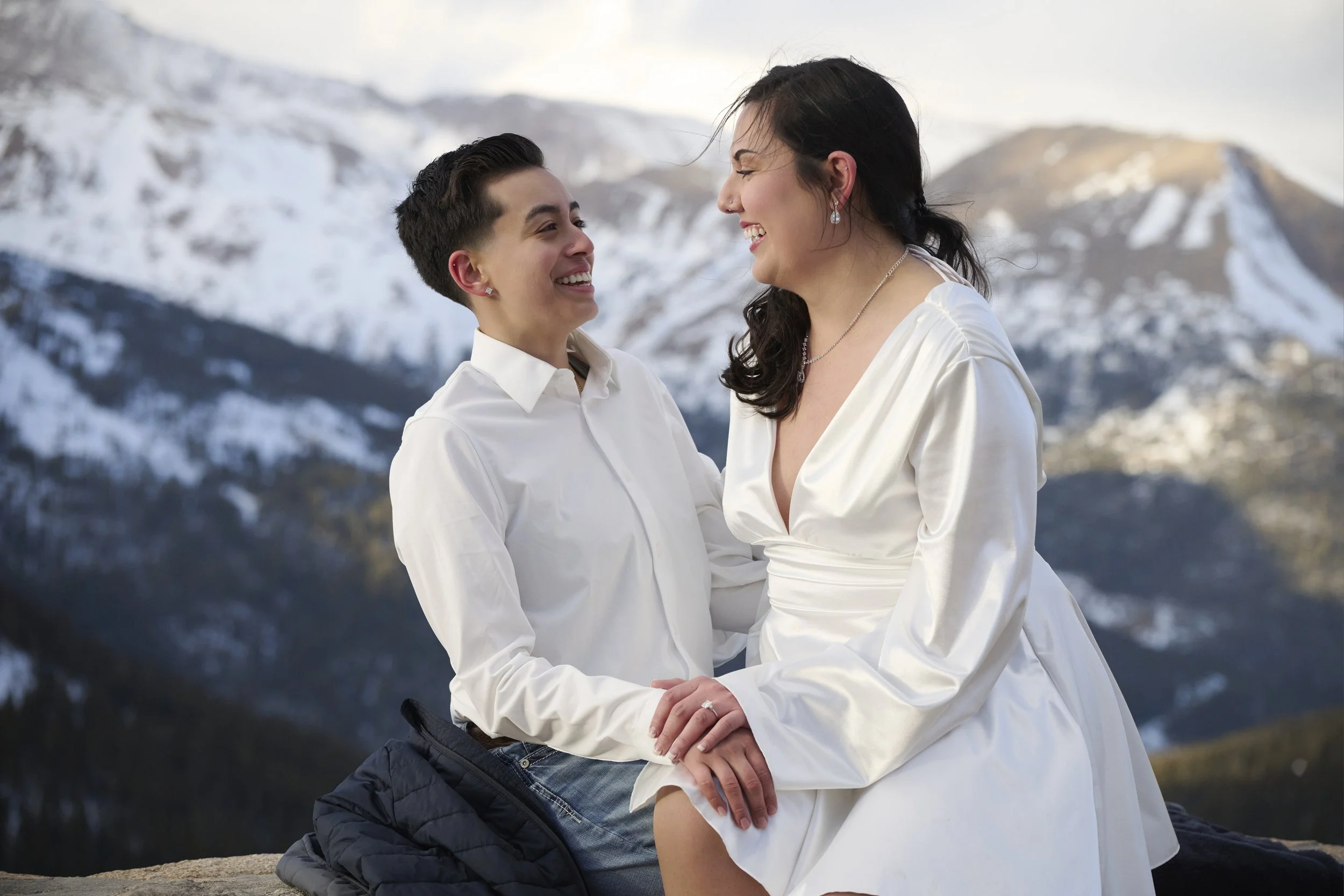 Two women smiling and holding hands, sitting outdoors with a snowy mountain landscape in the background.