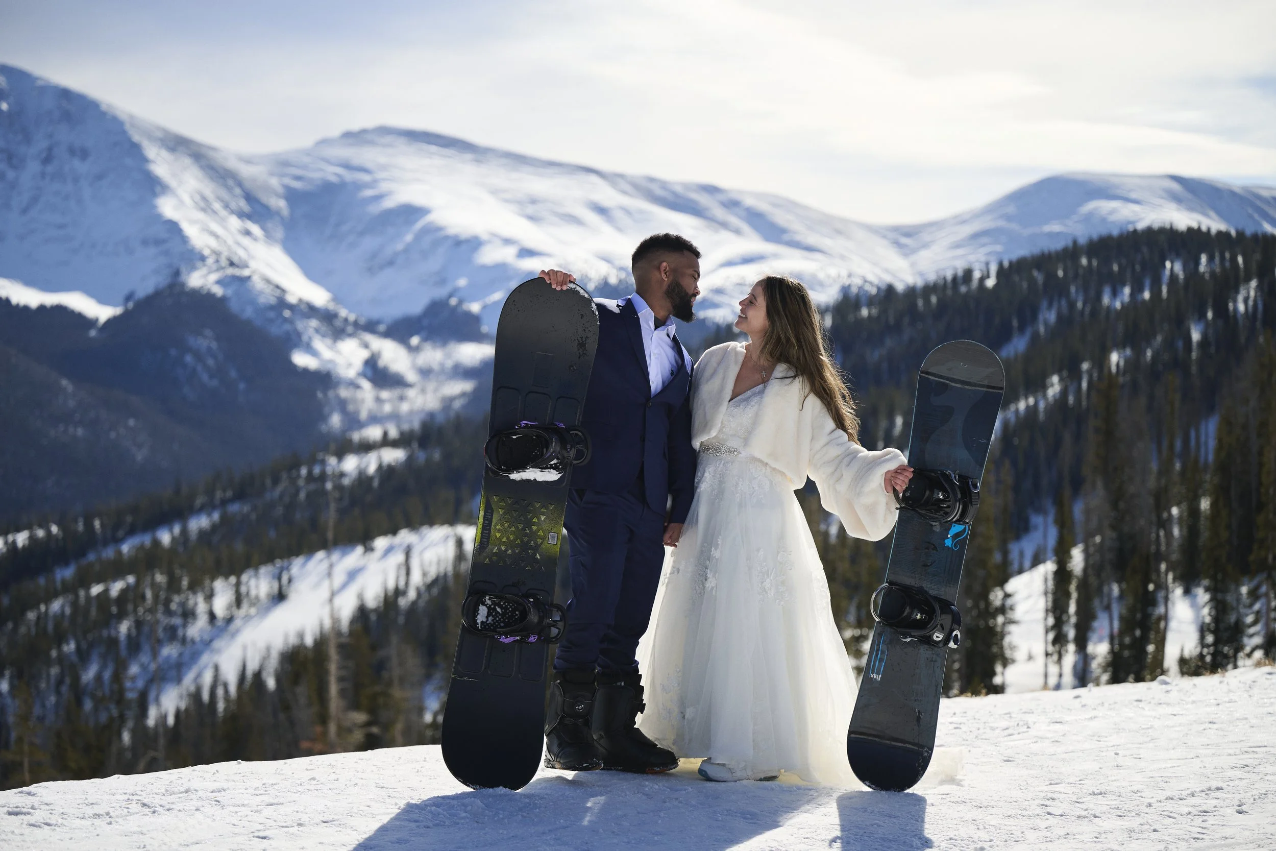 A couple in wedding attire standing in snowy mountains holding snowboard equipment, facing each other and smiling.