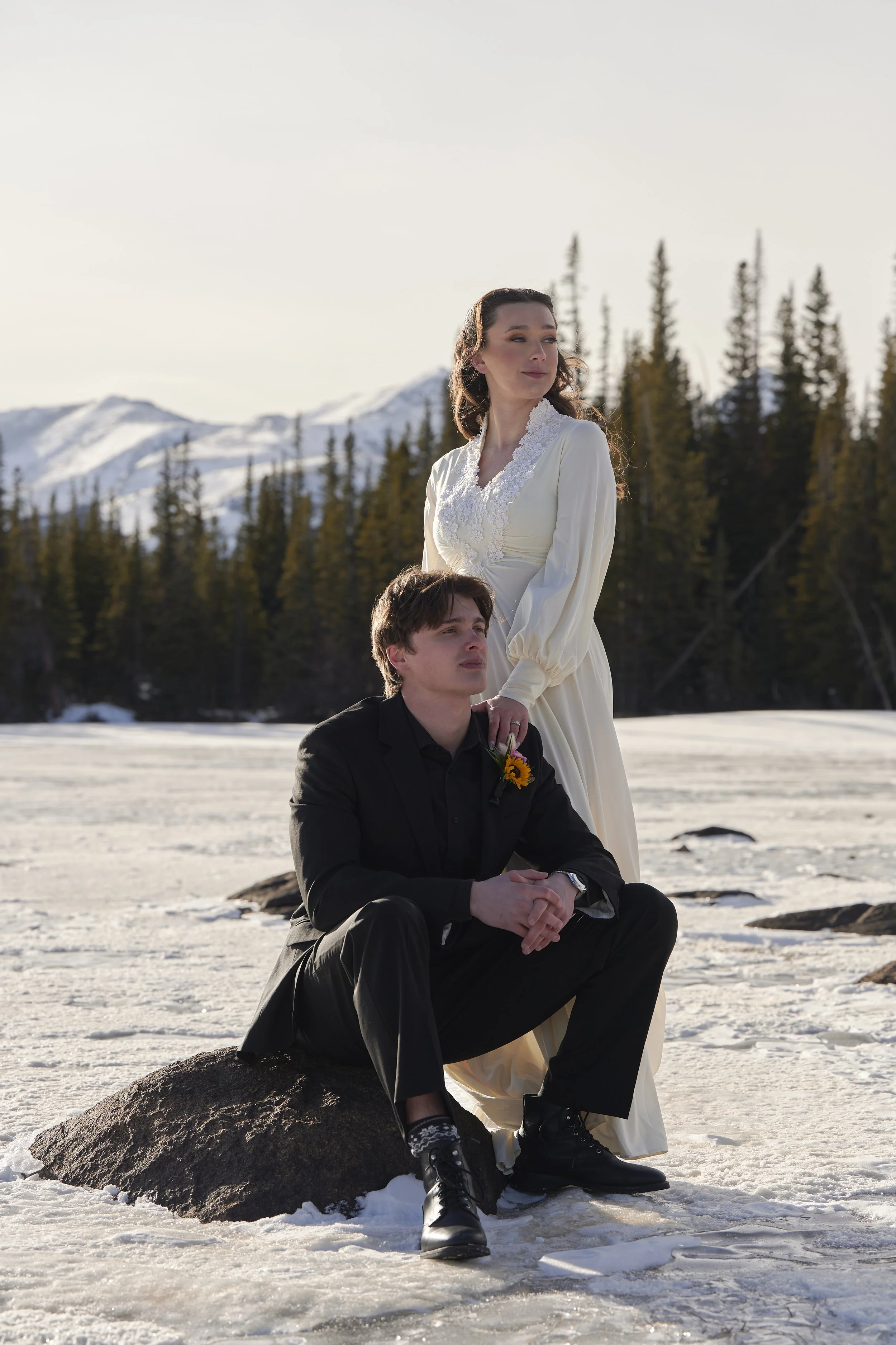 A woman in a white dress and a man in a black suit are outdoors on snow, with mountains and pine trees in the background. The woman stands, resting her hand on the man's shoulder as he sits on a rock.
