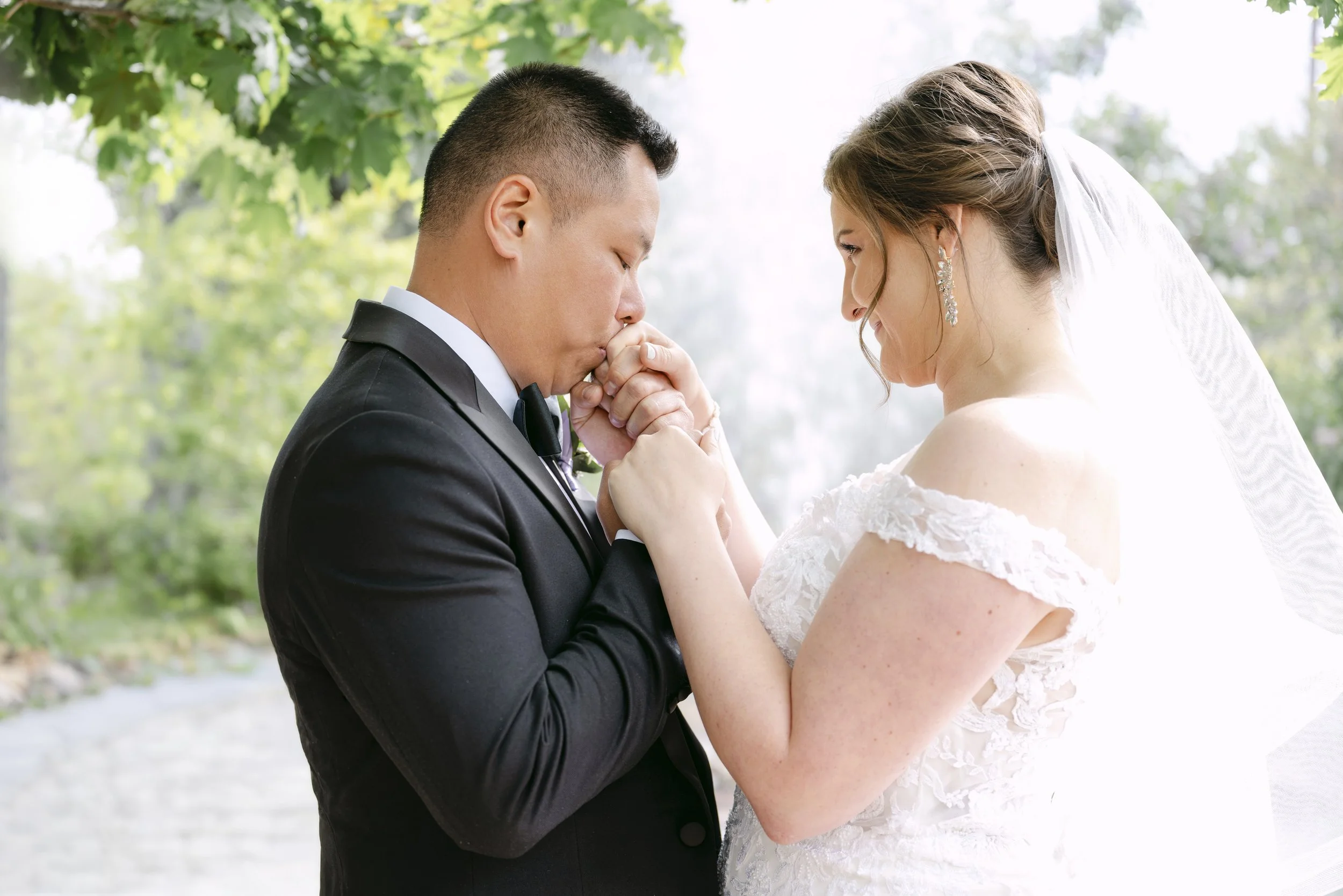 A bride and groom sharing a moment outside during their wedding, with the groom kissing the bride's hand as she looks at him.