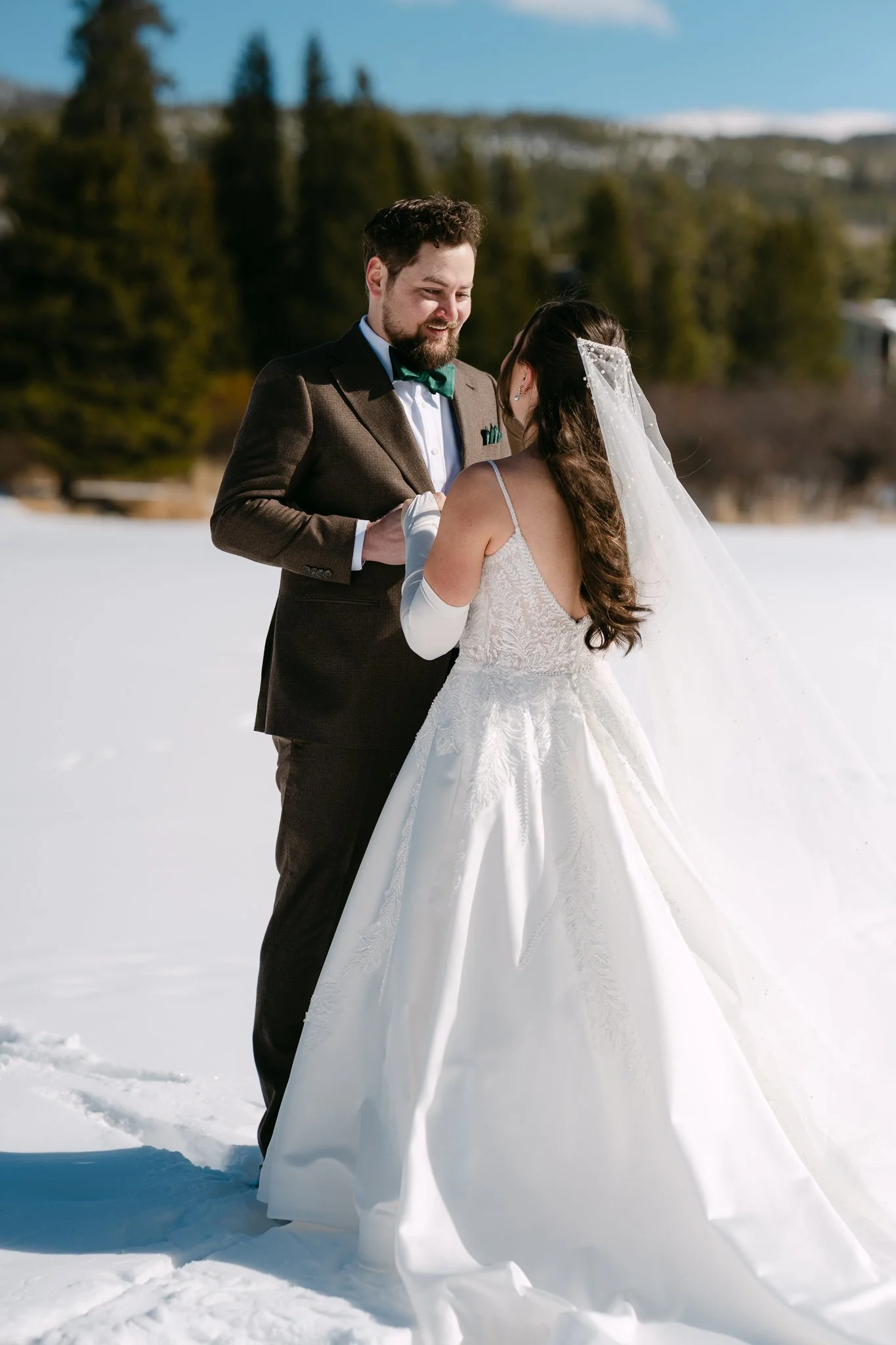 A bride and groom standing in snow outdoors, holding hands, sharing a moment during their wedding ceremony. The groom is wearing a brown suit with a green bow tie, and the bride is dressed in a white wedding gown with long gloves and a veil.