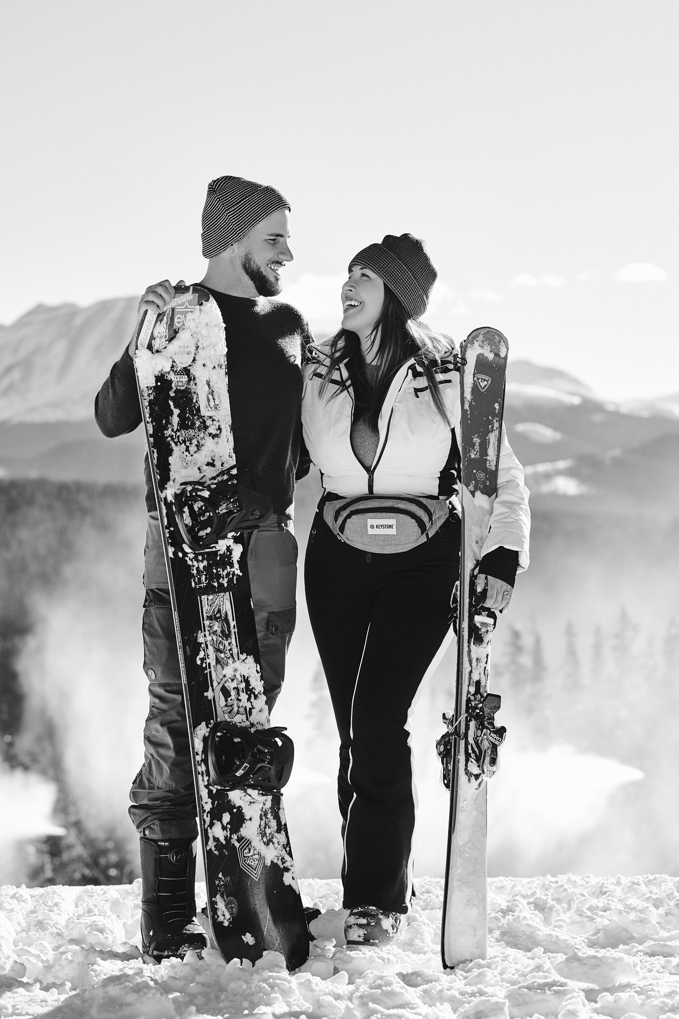 A man and woman in winter clothing holding snowboards, smiling at each other in a snowy mountain landscape.