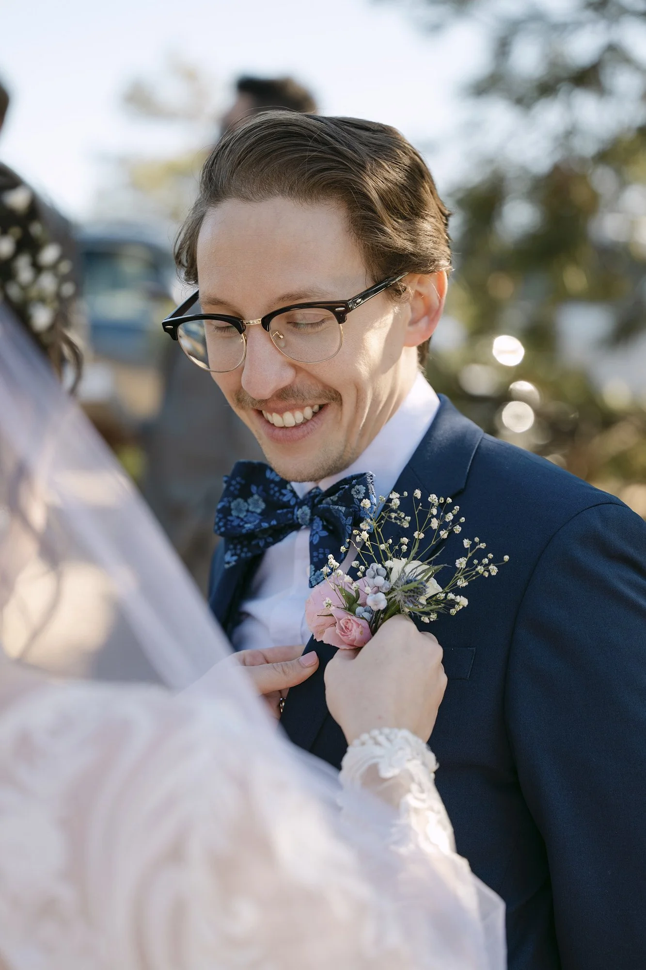 A smiling groom wearing glasses, a navy suit, and a blue bow tie with a floral boutonniere, on his wedding day.