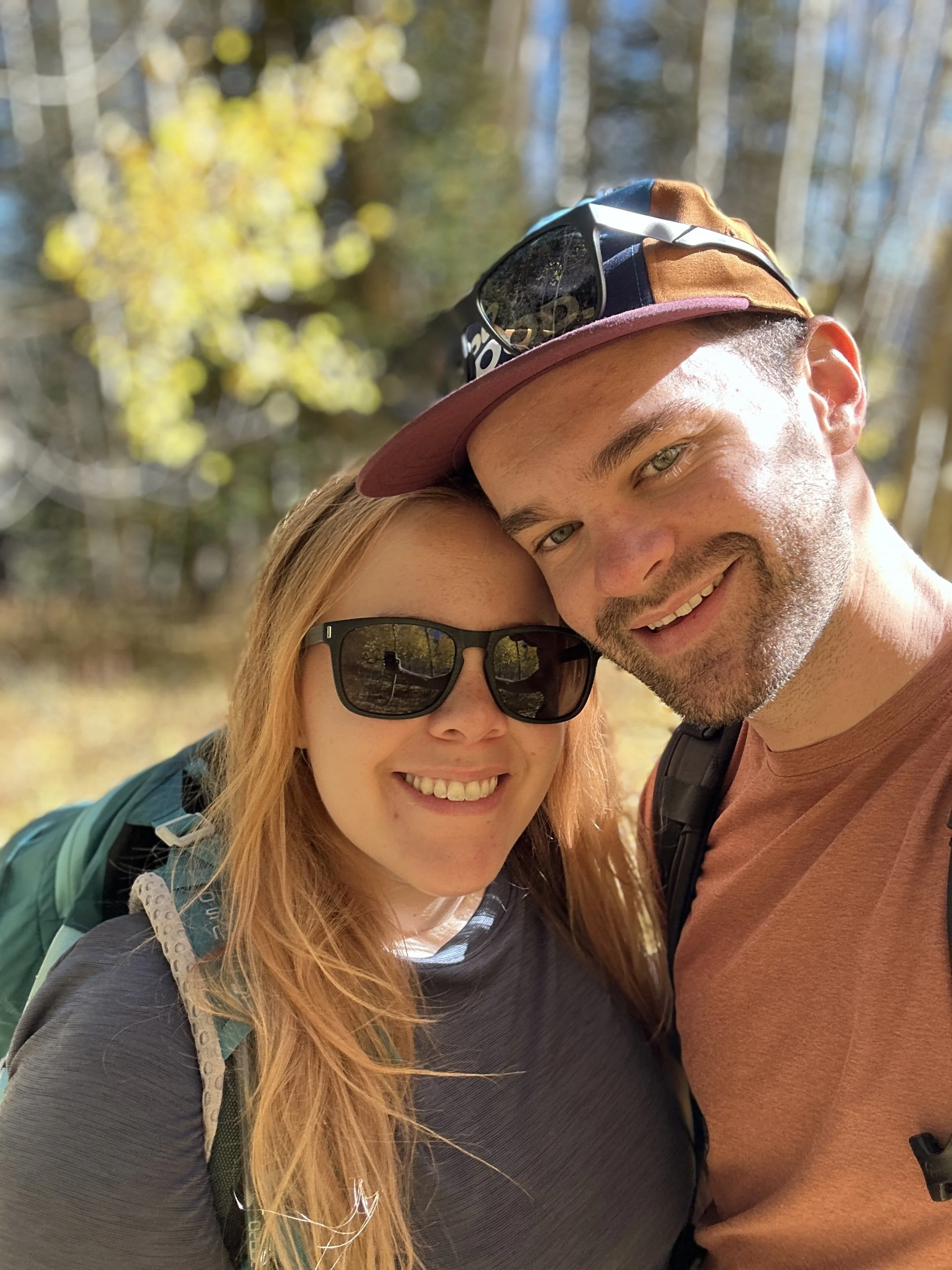 A smiling young woman and man taking a selfie outdoors in a wooded area during daytime.