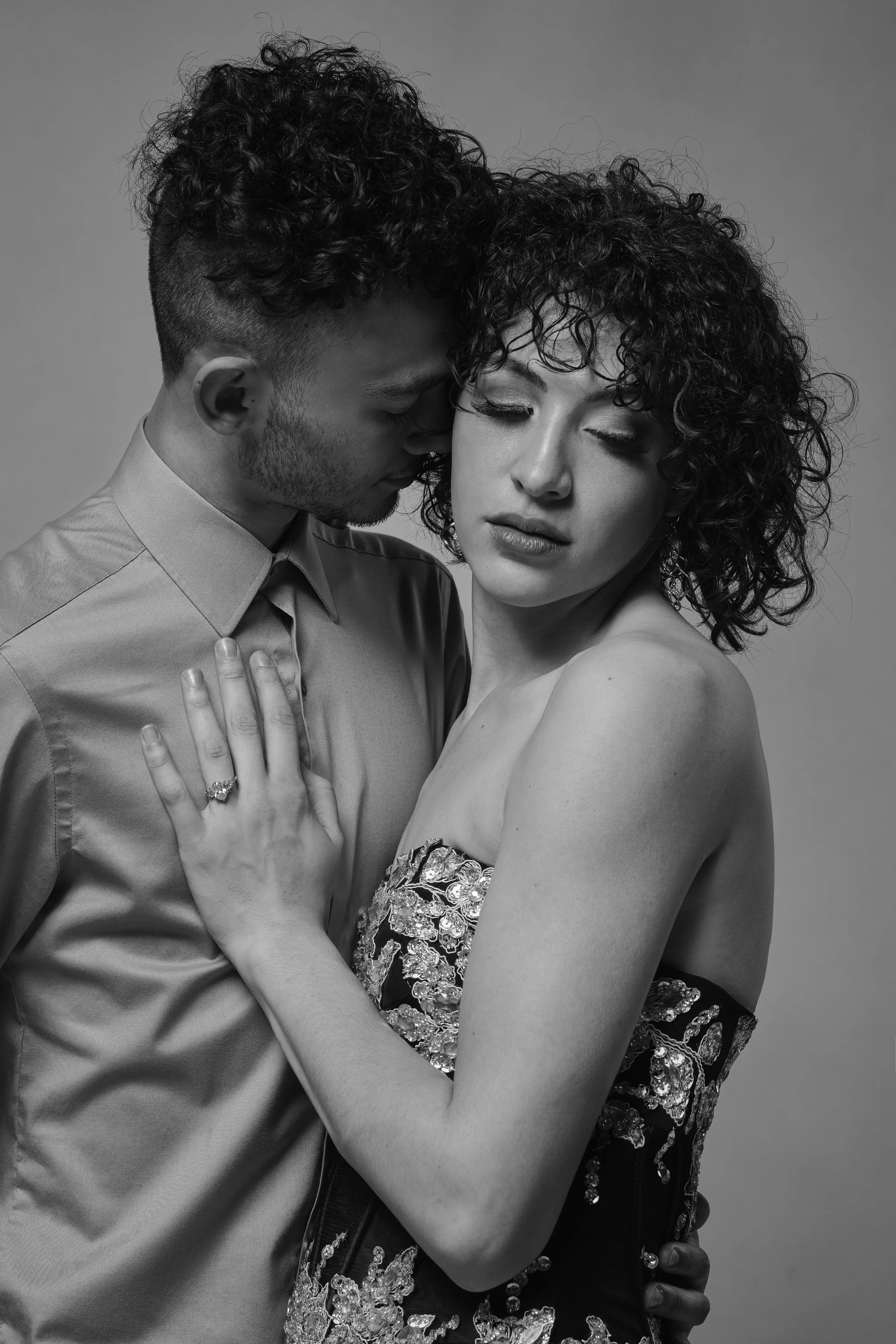 A black and white photo of a couple embracing closely. The man has curly hair and is wearing a button-up shirt, while the woman has curly hair and is wearing a strapless dress with floral embellishments. The woman has her hand on the man's chest, showing a ring, and their eyes are closed with tender expressions.