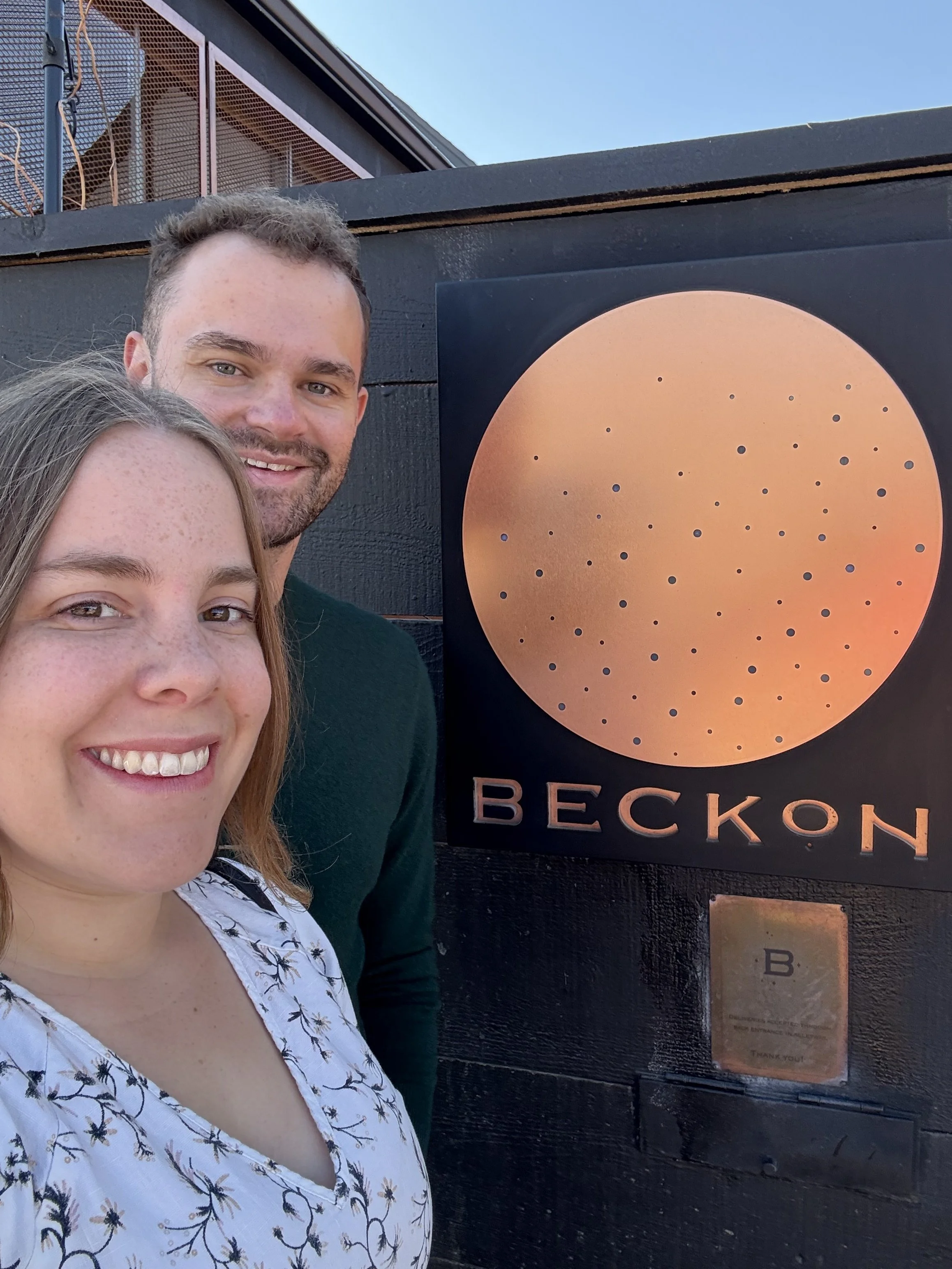 A woman and a man taking a selfie in front of a sign with a copper-colored circle and the word 'BECKON' in capital letters. The woman has long light brown hair and freckles, and the man has short brown hair and a beard. They are smiling and standing outdoors on a clear day.