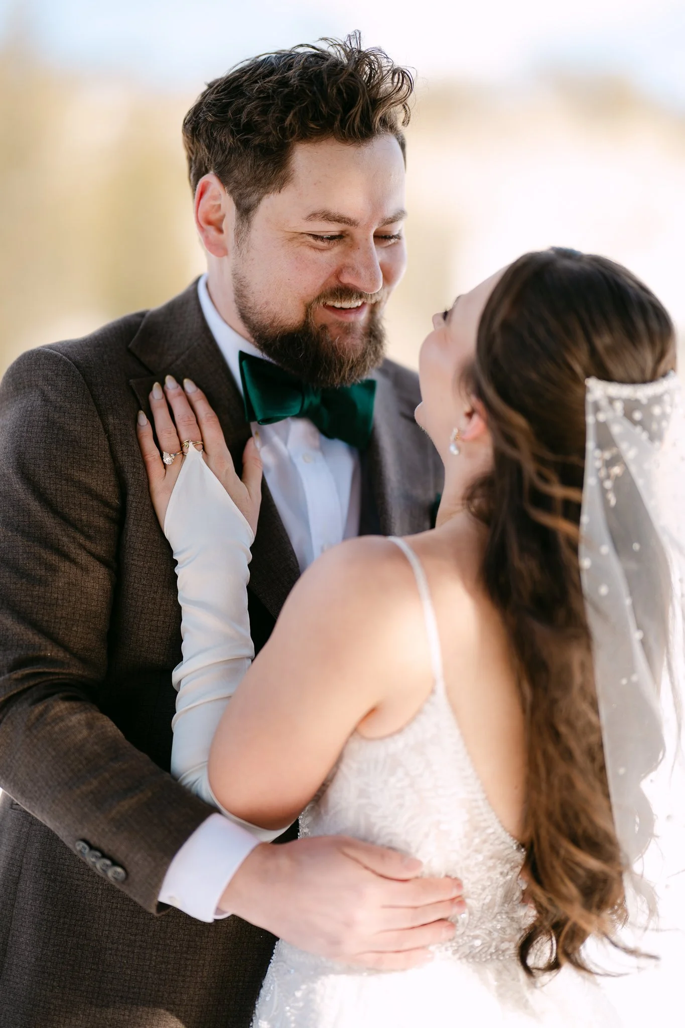 A bride and groom sharing a close moment on their wedding day. The groom, wearing a dark suit and green bow tie, smiles as he looks at the bride. The bride, in a lace wedding dress and long gloves, looks up at the groom with a joyful expression, her hand resting on his shoulder.
