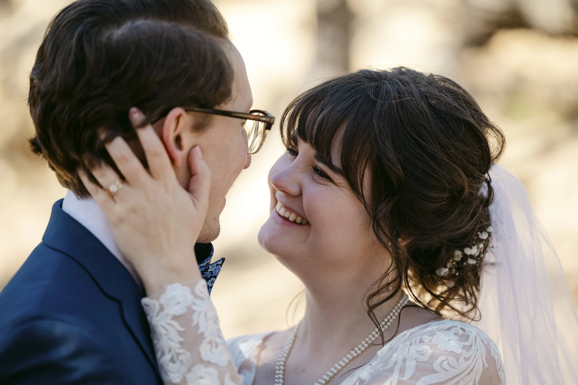 A bride and groom looking at each other, smiling, with their noses almost touching. The bride is holding the groom's face with her hand and wearing a wedding dress, veil, and pearls. The groom is dressed in a navy suit and glasses.