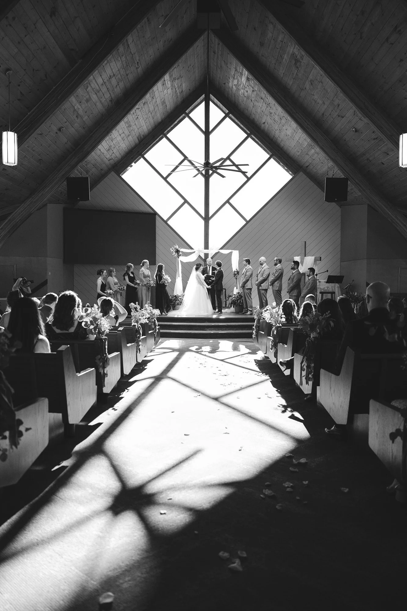 A black and white photo of a wedding ceremony inside a church with a high, wooden, A-frame ceiling and large glass window behind the altar. The bride and groom stand at the altar exchanging vows, surrounded by their wedding party, with guests seated 