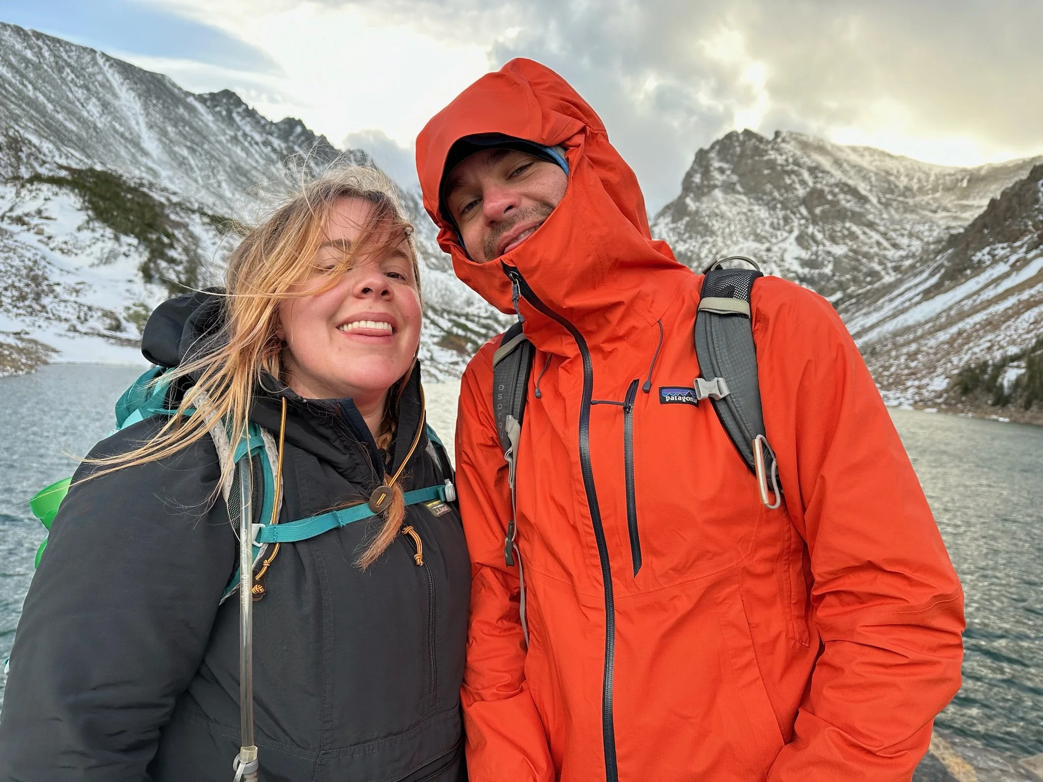 A smiling woman and a man in an orange jacket taking a selfie outdoors near a lake with snow-covered mountains in the background.