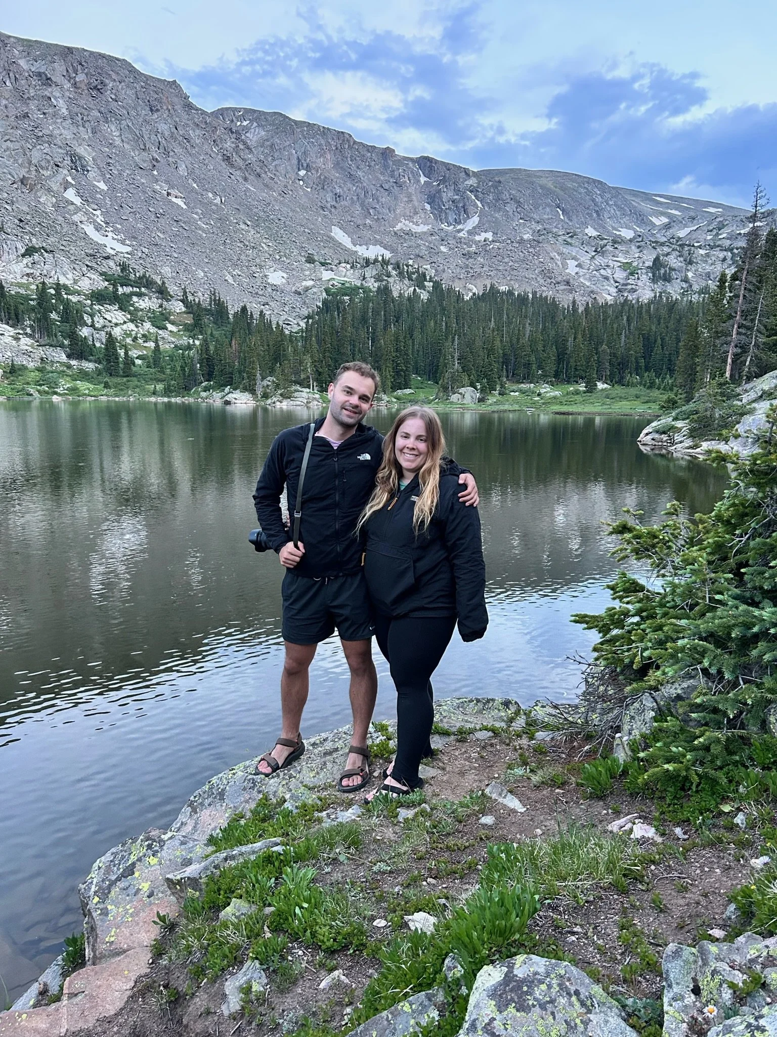 A young man and woman standing close together on a rock by a lake in a mountainous area with pine trees and snow patches, smiling at the camera.