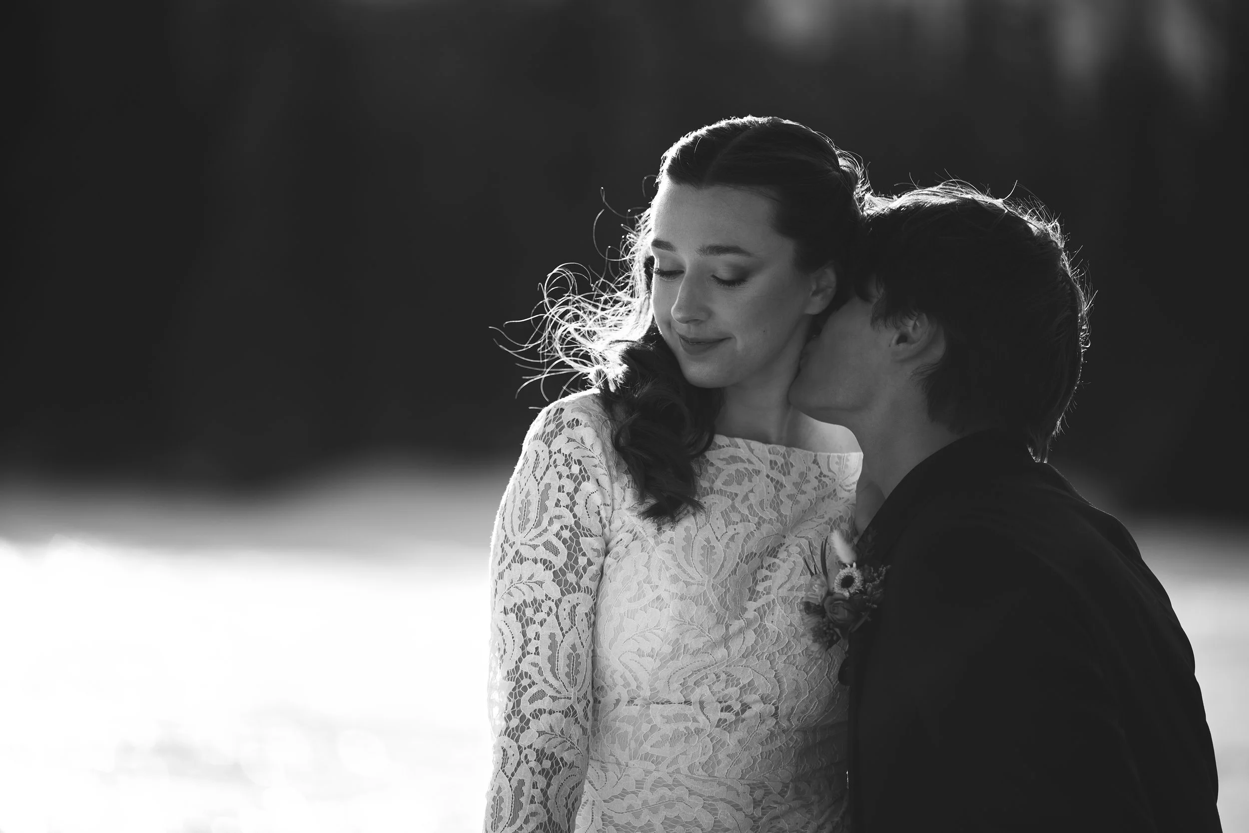 Black and white photo of a woman in a lace wedding dress and a man, with the man kissing the woman's cheek, outdoors in a natural setting.