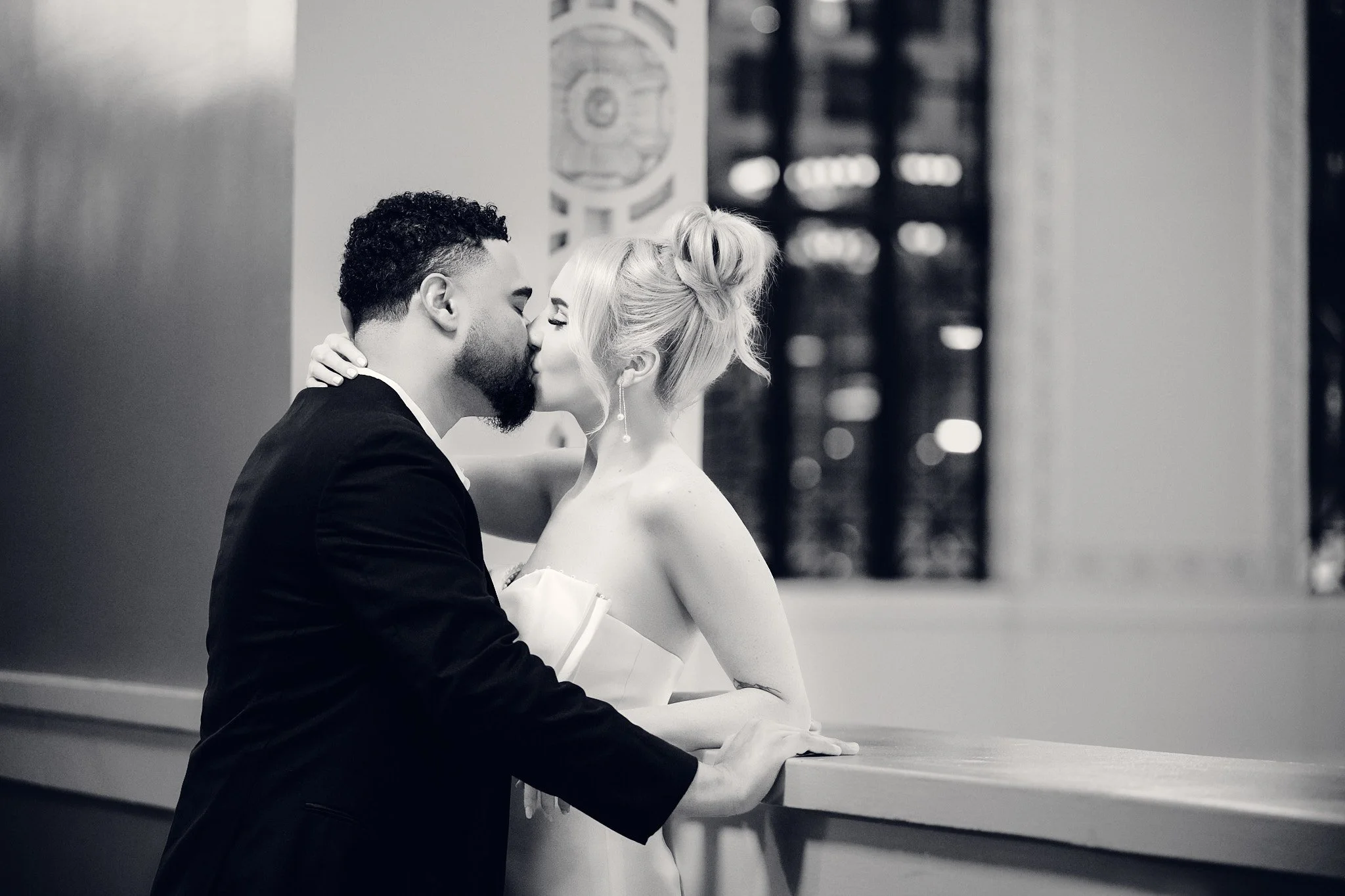 A couple sharing a kiss, with the man in a suit and the woman in a strapless dress, standing at a bar or counter inside a building.